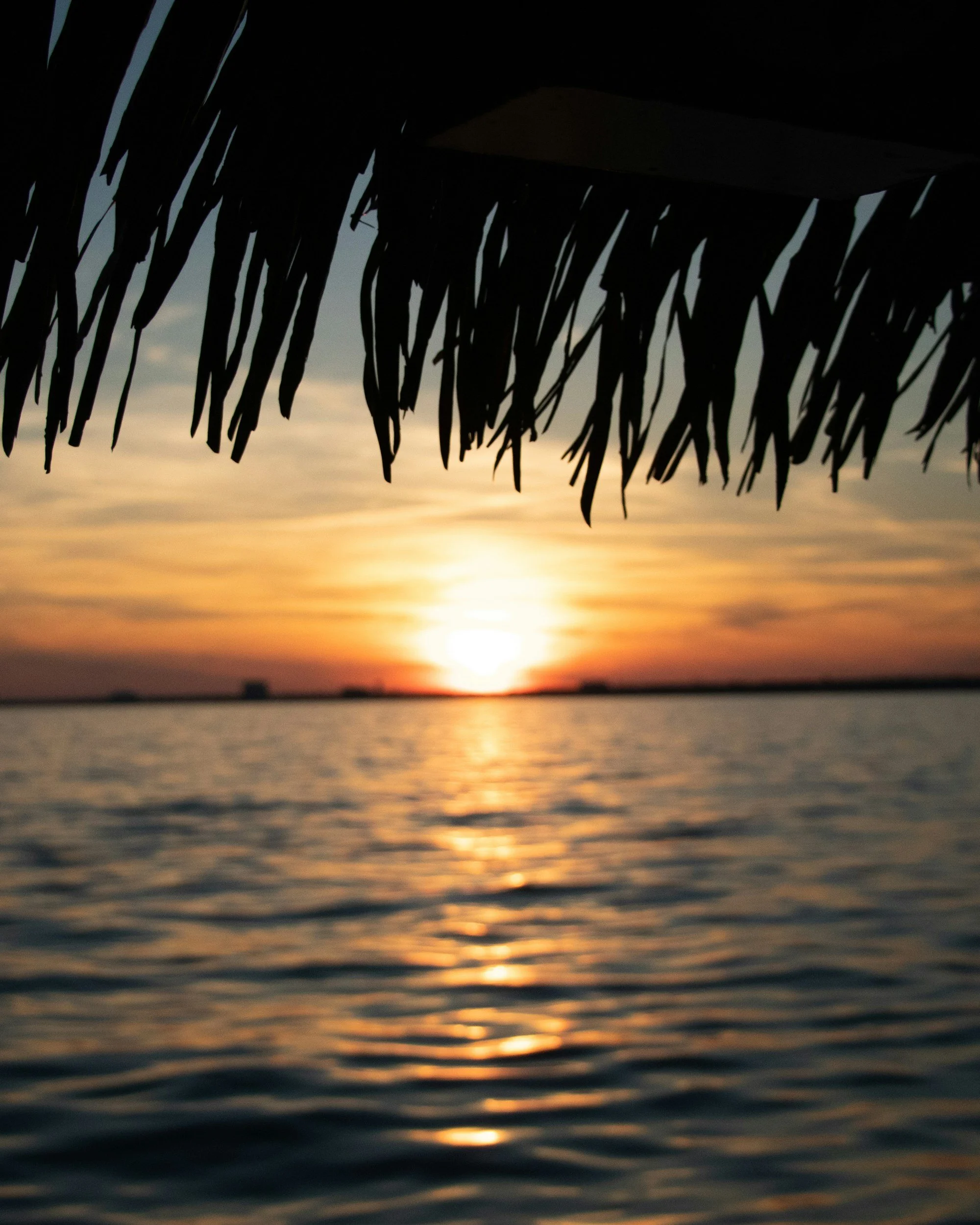 Silhouette of a thatched roof over water at sunset with a colorful, orange, and yellow sky in the background.