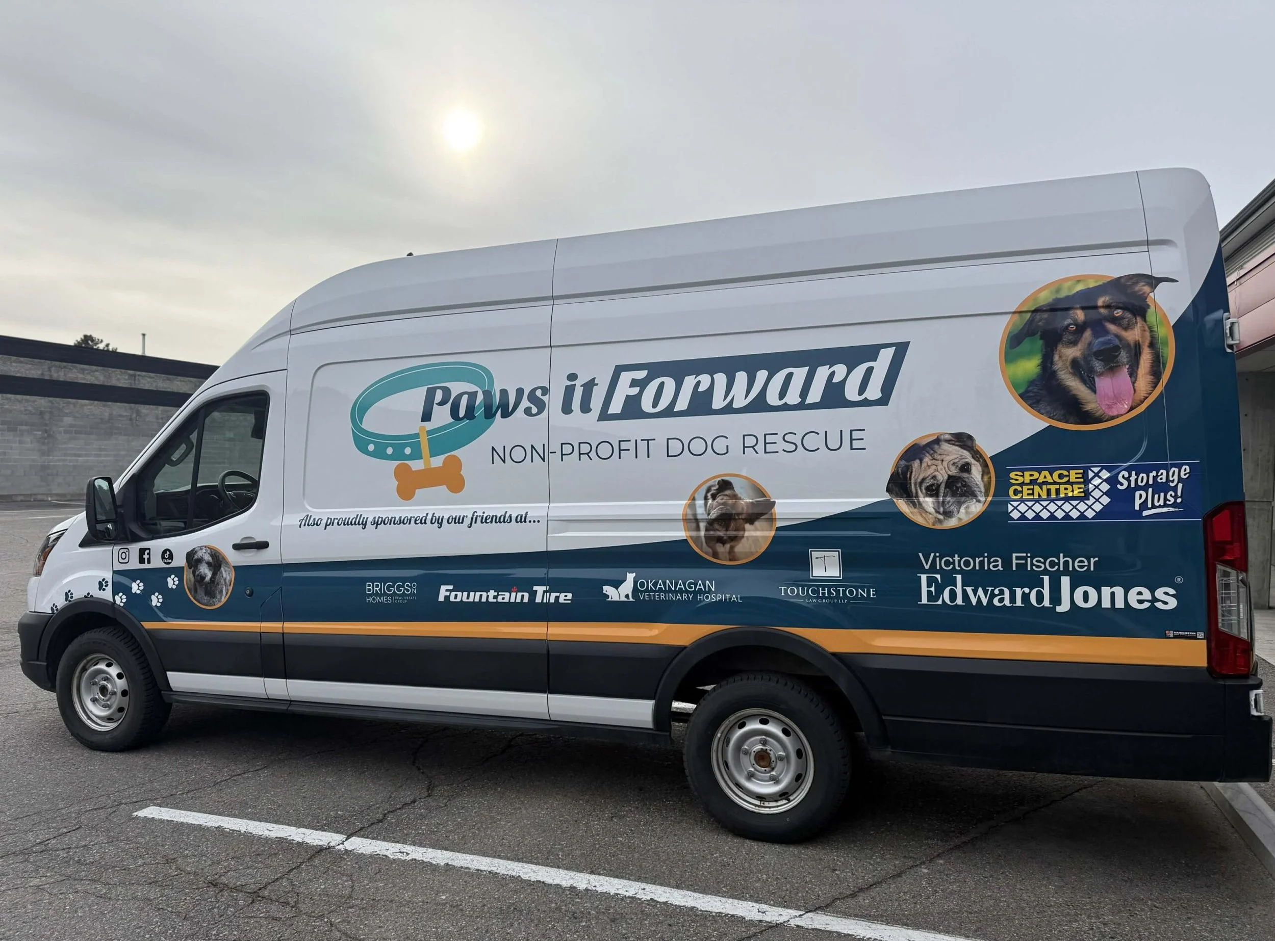 A white van parked in a parking lot with a graphic design promoting a dog rescue organization called Paws it Forward. The van features a large image of a happy, black and tan dog with its tongue out, and multiple smaller images of different dog breeds. The van displays logos of sponsors and partners, including Edward Jones, Fountain Tire, and Okanagan Veterinary Hospital. The sky is overcast, and the background shows a building and some trees.