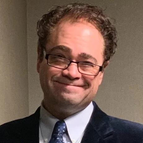 Portrait of a man with curly hair, glasses, and a suit, smiling in front of a plain wall.