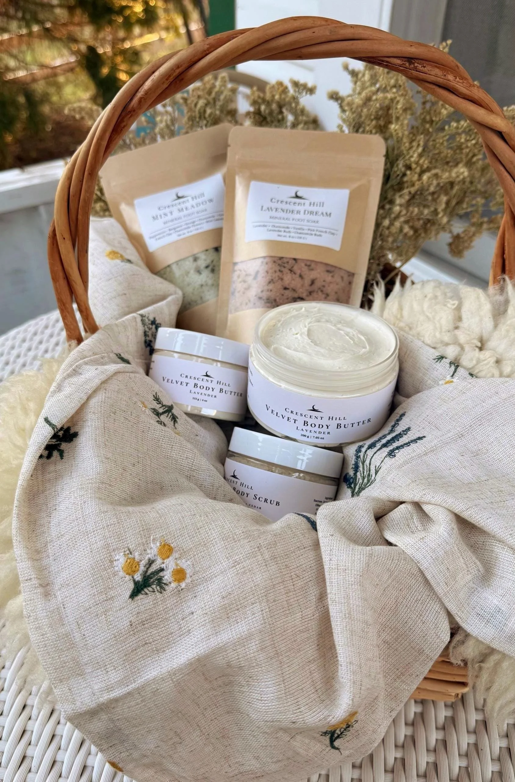 A wicker basket containing various self-care products including lavender scented body butter, body scrub, and bath salts, with dried flowers in the background.