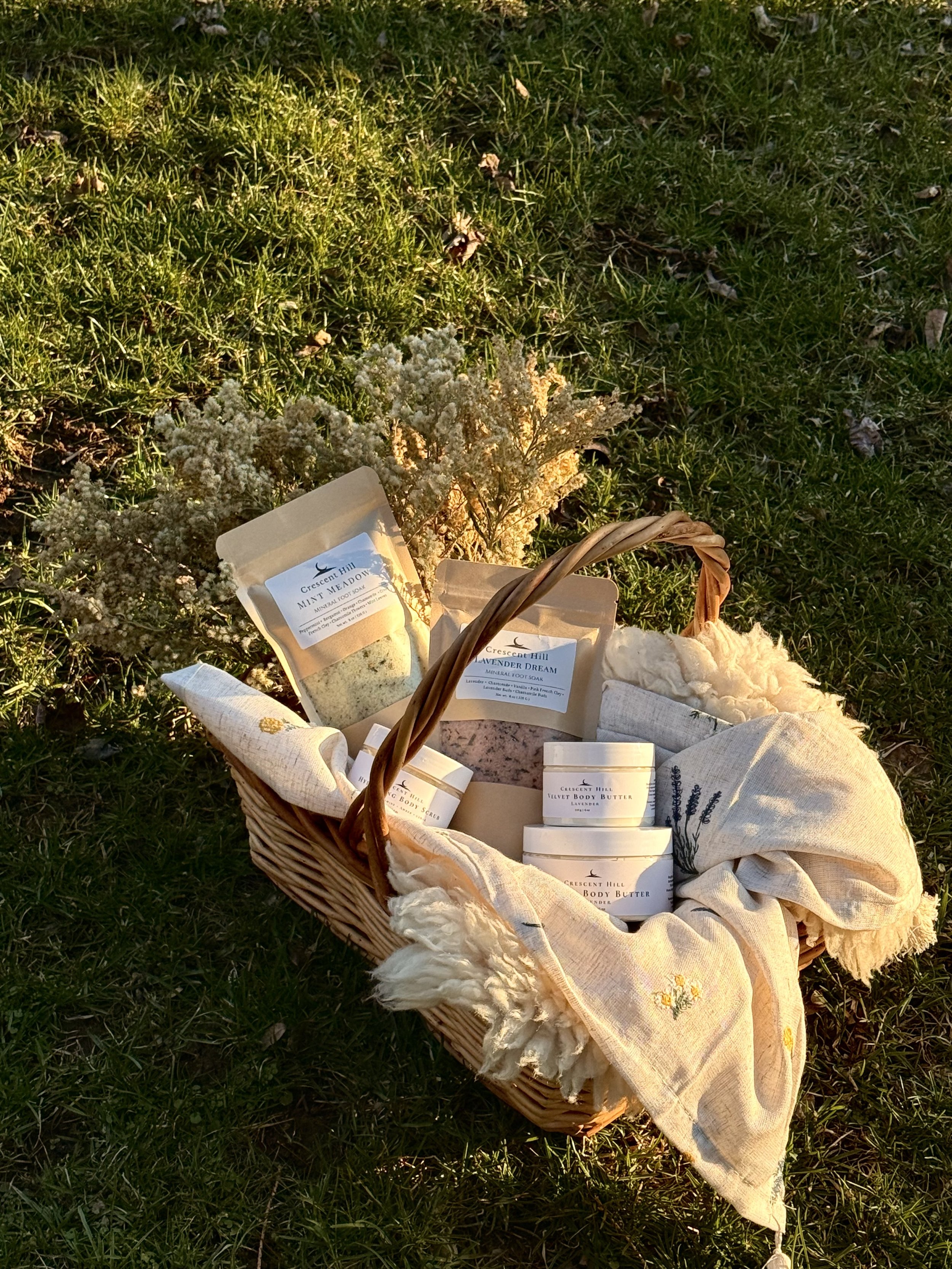 A wicker basket on the grass filled with skincare products, including body butter and mineral foot soak, with a cozy blanket.
