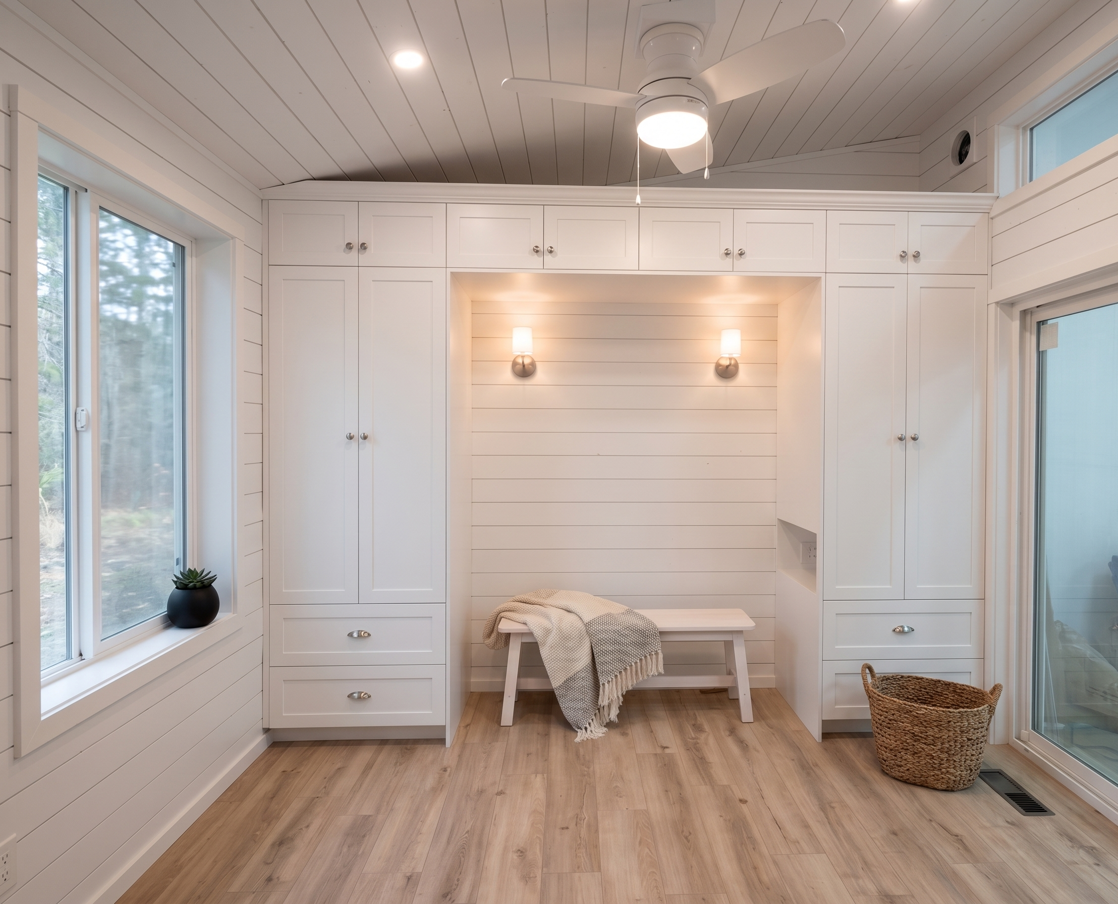 A bright, white, and cozy enclosed porch with large windows, built-in cabinets, and light wood flooring. There is a small white bench with a blanket, a potted succulent, and a wicker basket in the space. Two wall-mounted lights and a ceiling fan prov