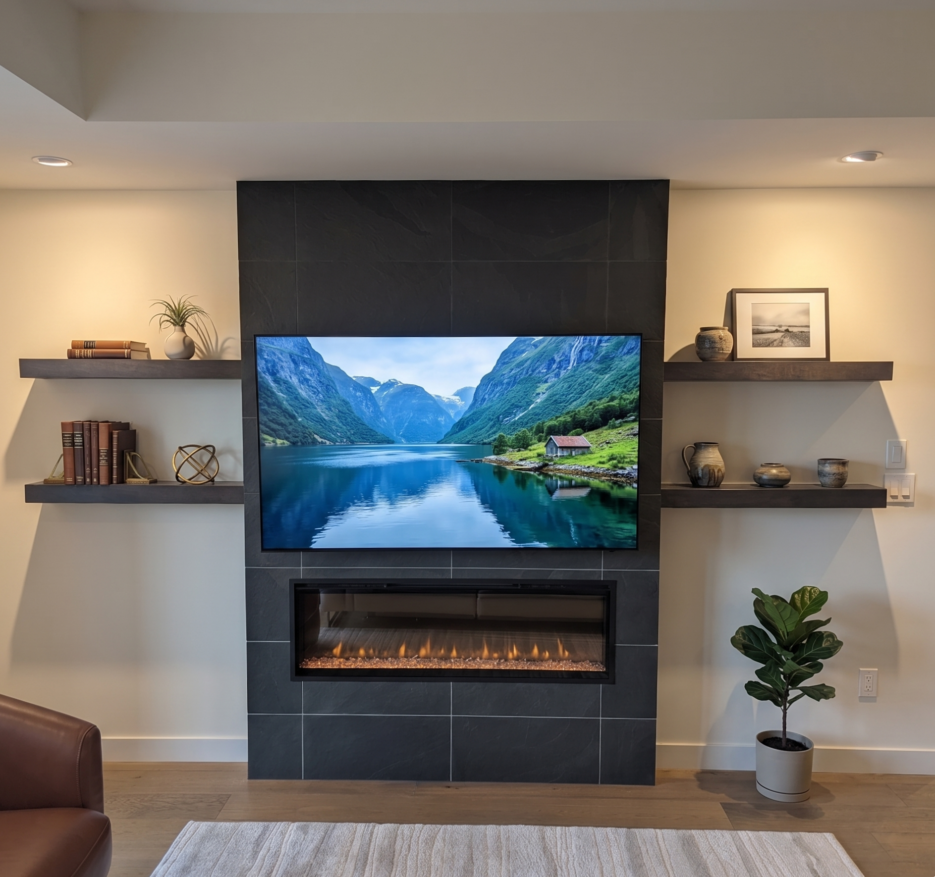 Modern living room with a mounted flat-screen TV displaying a scenic landscape, a gas fireplace beneath, and beige walls with dark shelves holding books, decorative vases, and framed artwork; a potted plant on the right, hardwood flooring, and a beig