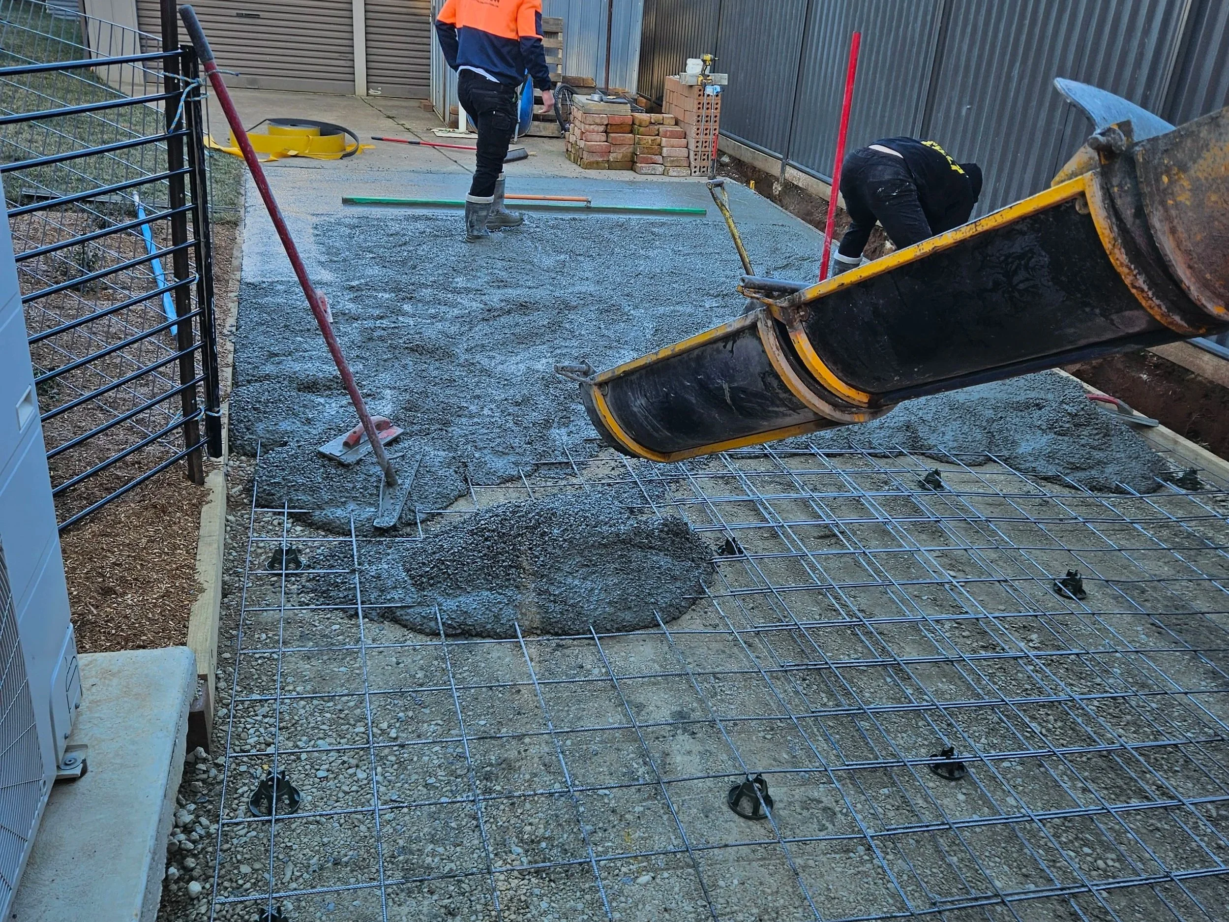 Construction workers are preparing a concrete slab on a ground surface, with rebar grid and tools visible, in an outdoor area enclosed by fences and nearby stacks of bricks.