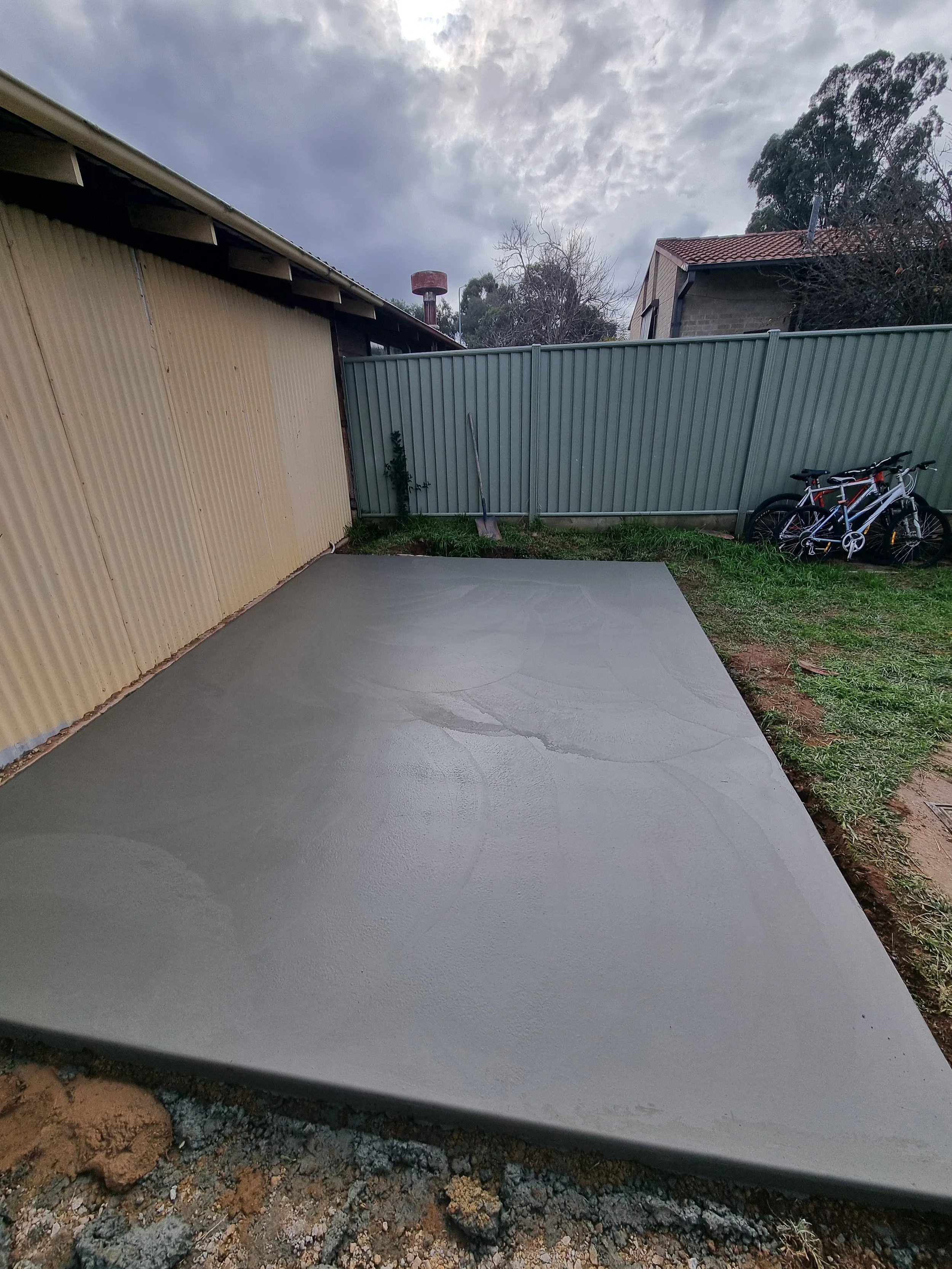 Freshly poured concrete slab in a backyard with grass, a metal fence, and two bikes leaning against it. Overcast sky with clouds.