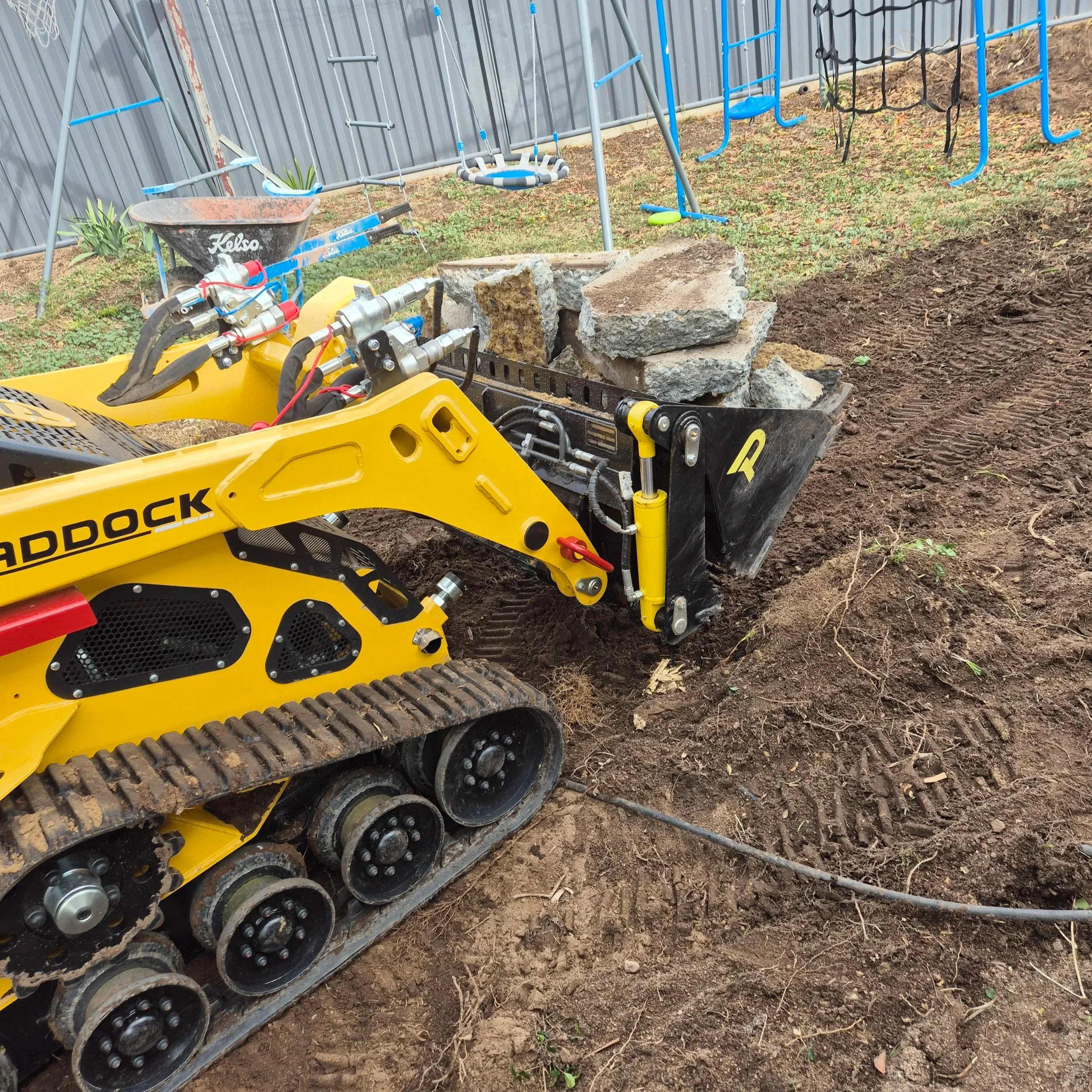 Construction site with a small tracked excavator lifting large rocks, a metal fence in the background, and a playset with swings and a climbing structure nearby.