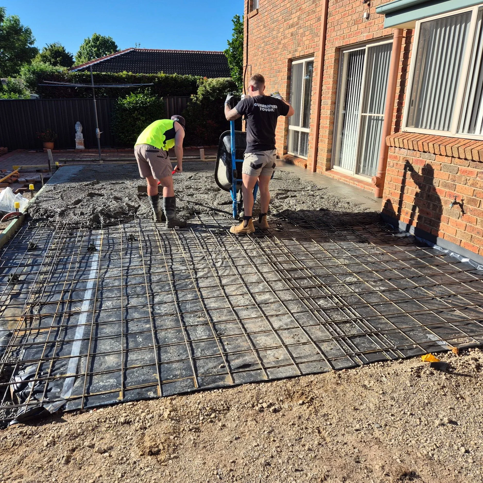 Two workers leveling and preparing a concrete slab foundation in a backyard, with a brick house and green trees in the background.
