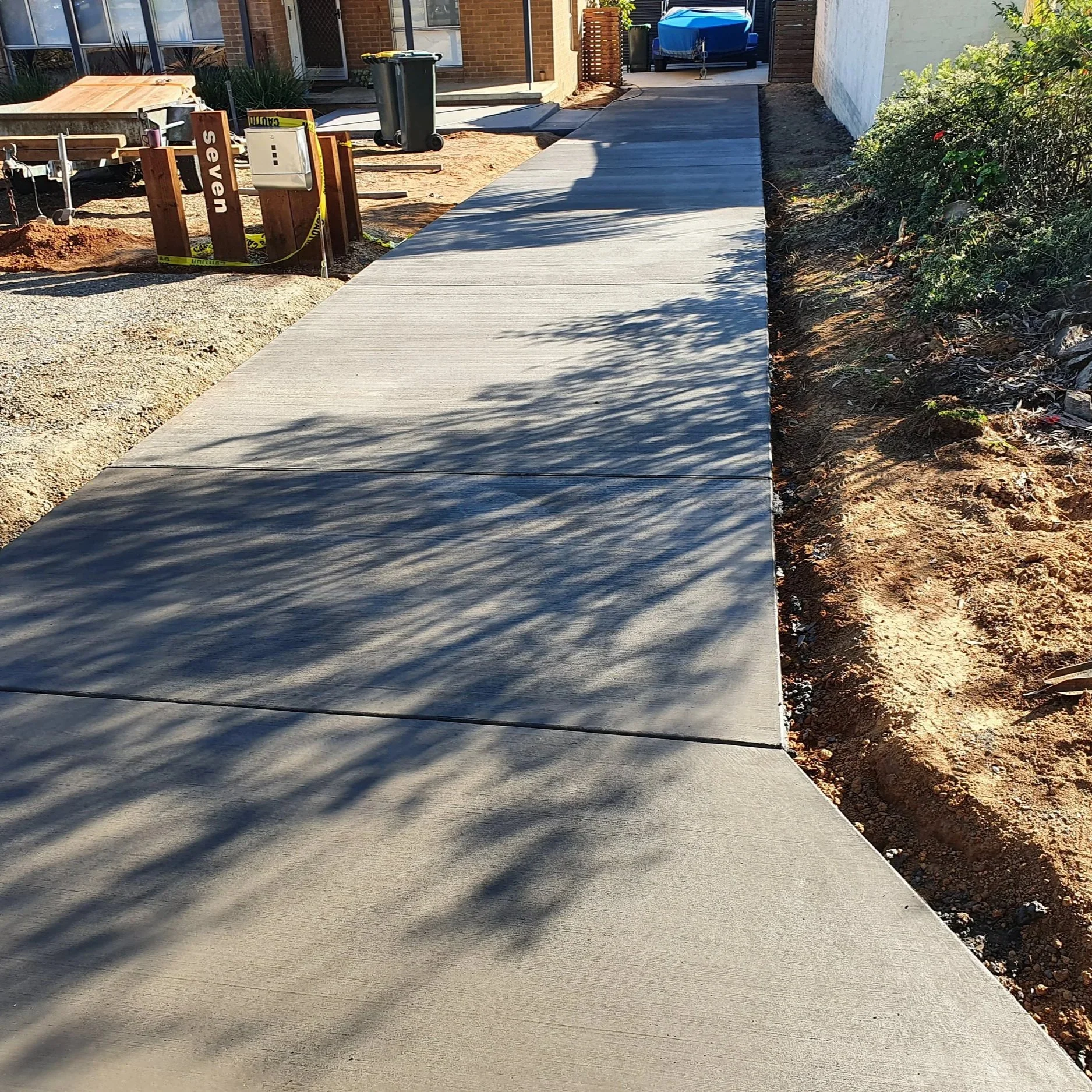 Newly poured concrete sidewalk with shadows from nearby trees on a sunny day, adjacent to a dirt and gravel area, leading to a house and yard with plants and outdoor furniture.