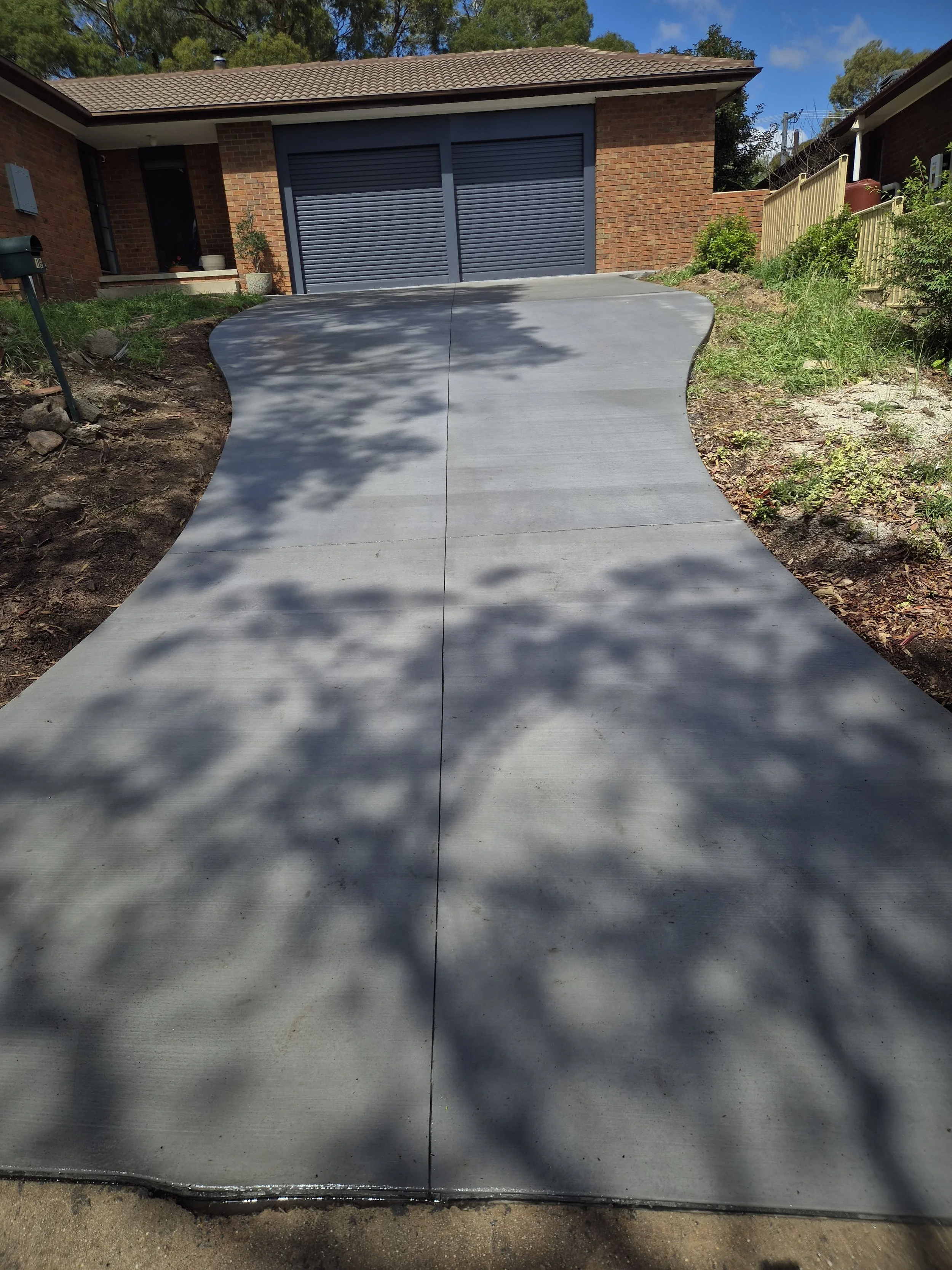 Newly poured concrete driveway leading up to a garage with blue shutters at a brick house. Shade patterns from nearby trees are visible on the concrete surface.