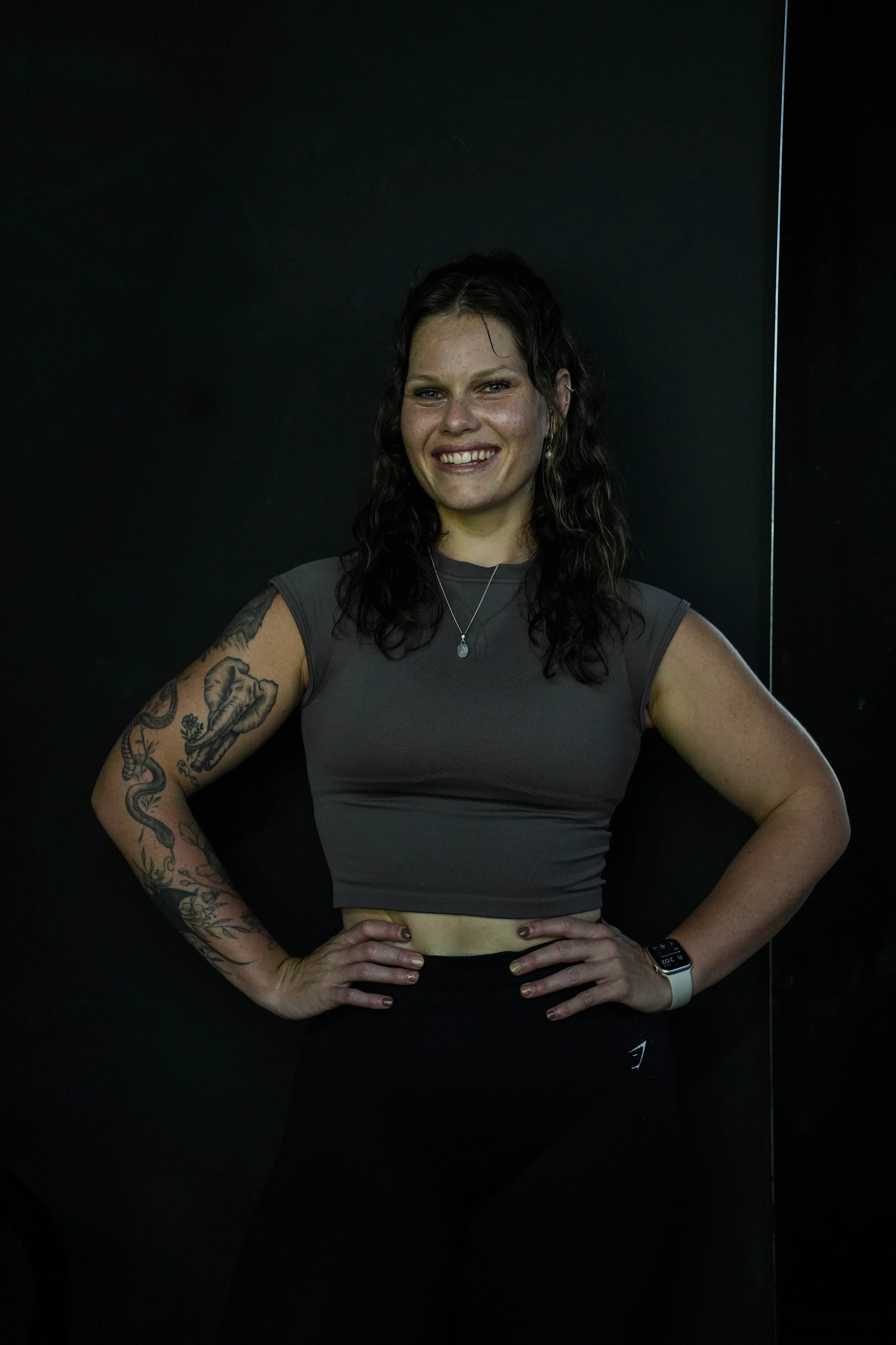 A woman with curly dark hair, tattoos on her right arm, wearing a dark gray sleeveless top, smiling, standing with hands on hips against a black background.
