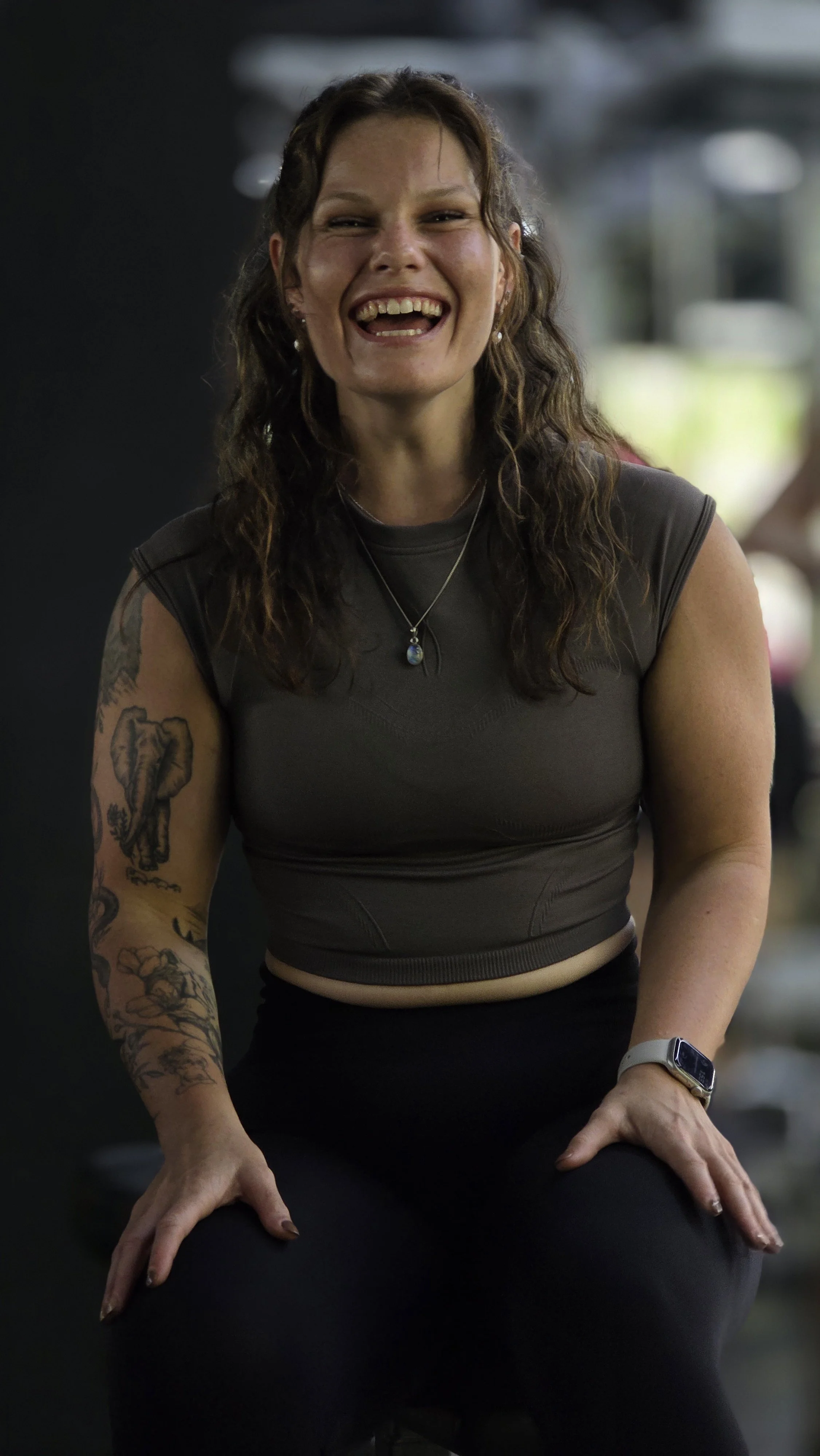 A woman with curly brown hair, tattoos on her arms, and wearing a gray athletic top and black pants, sitting in a fitness or yoga pose in a gym or studio. She is smiling and looking directly at the camera.