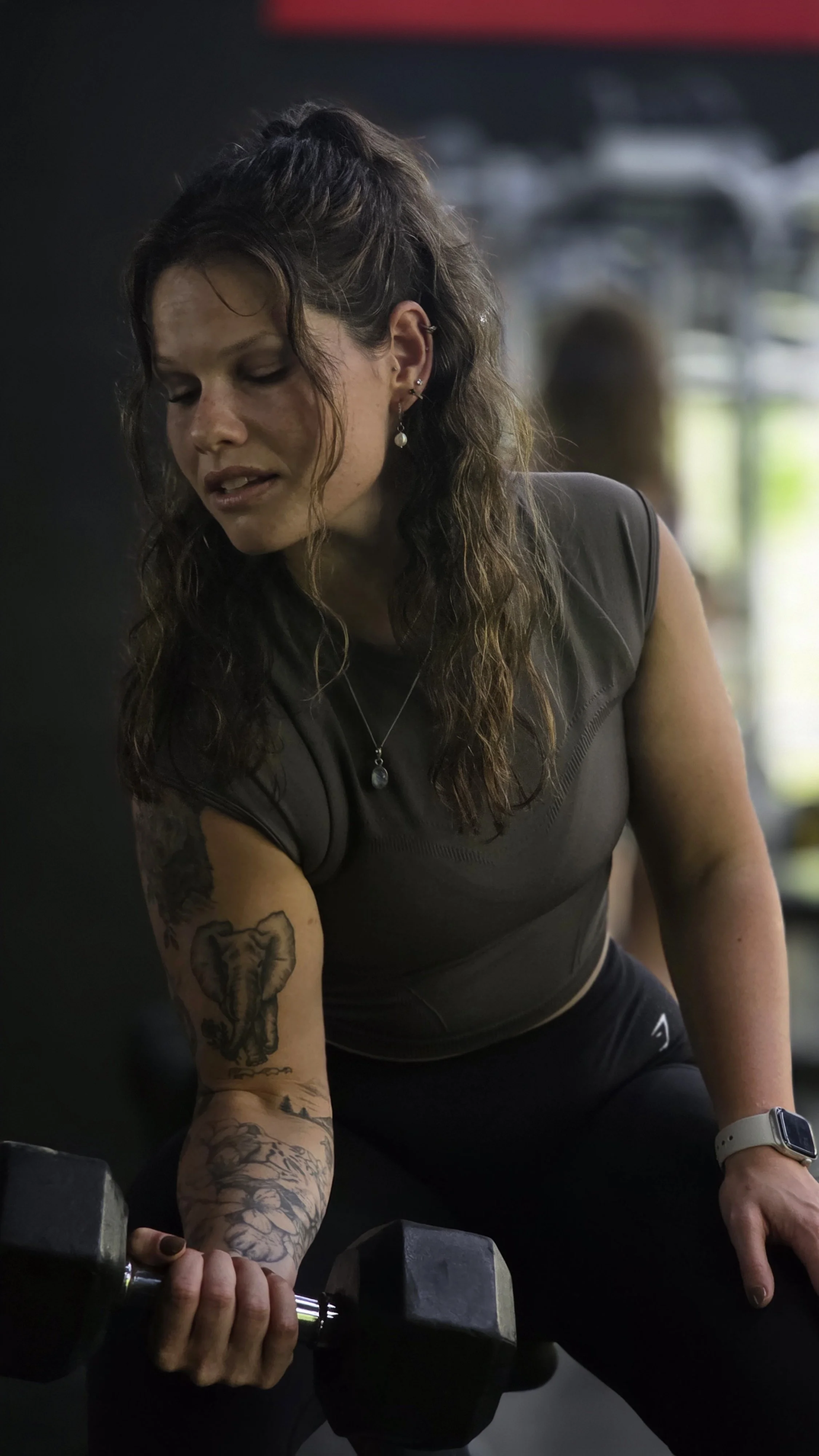 A woman with curly brown hair and tattoos on her right arm is lifting a black dumbbell in a gym. She is wearing a dark workout top and a watch, with a focused expression.