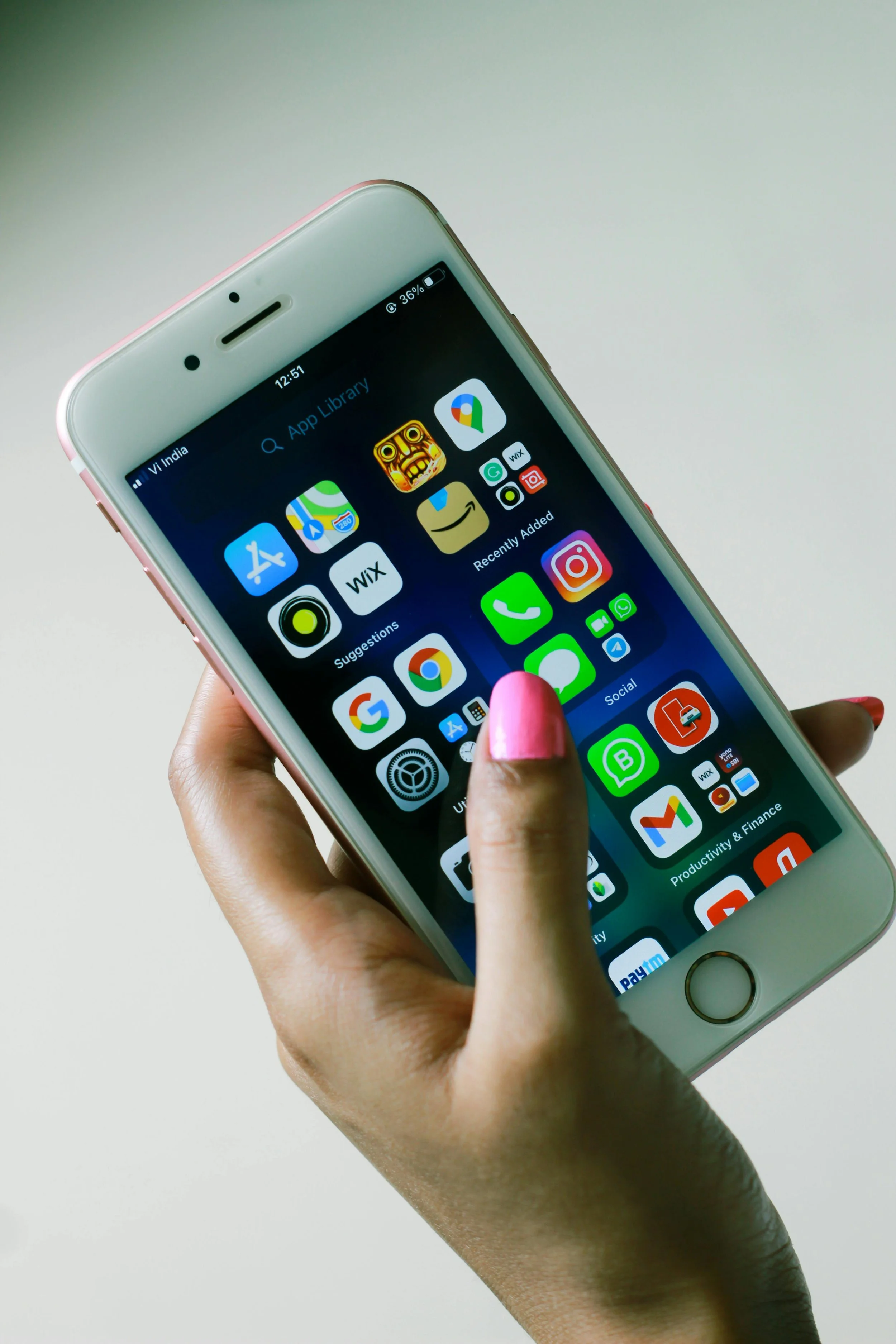 Close-up of a person's hand holding a smartphone with various app icons on the screen. The person has pink nail polish and a dark skin tone. The background is plain and light-colored.