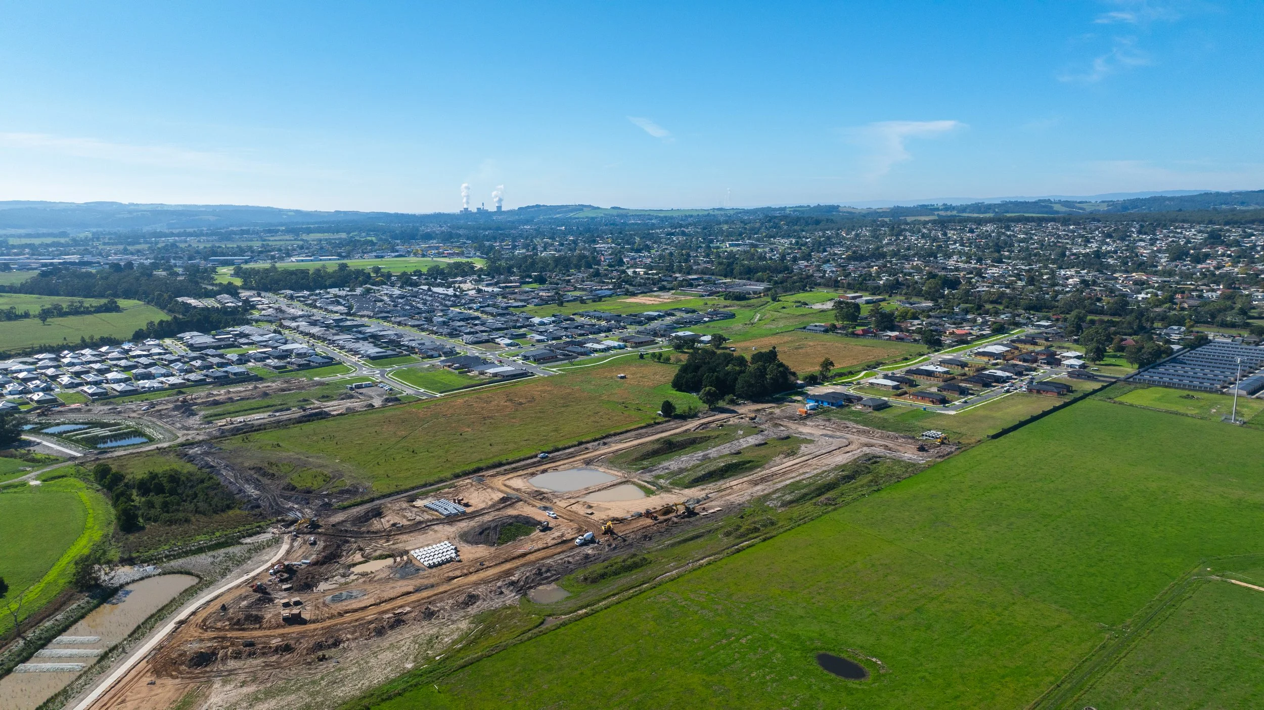 Aerial view of a developing suburban area with residential neighborhoods, green fields, and ongoing construction, under a bright blue sky.