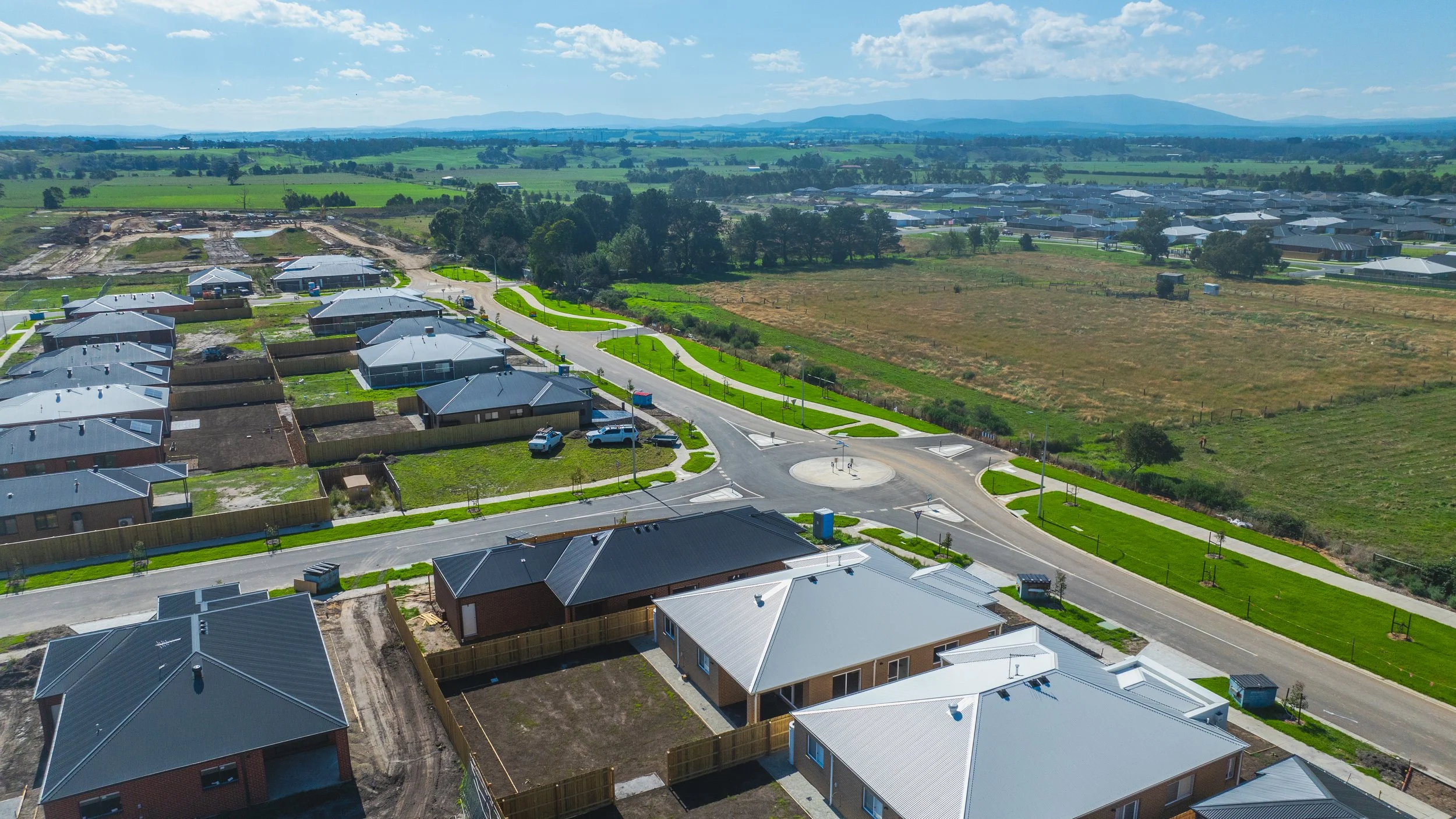 Aerial view of a suburban neighborhood with new houses, streets, and green lawns, with more houses and open fields in the distance under a partly cloudy sky.