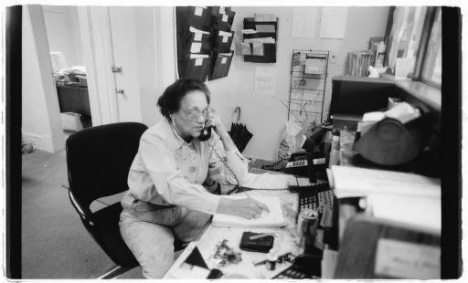 An elderly woman sitting at a cluttered desk, using a landline phone and working with papers and a calculator in an office.
