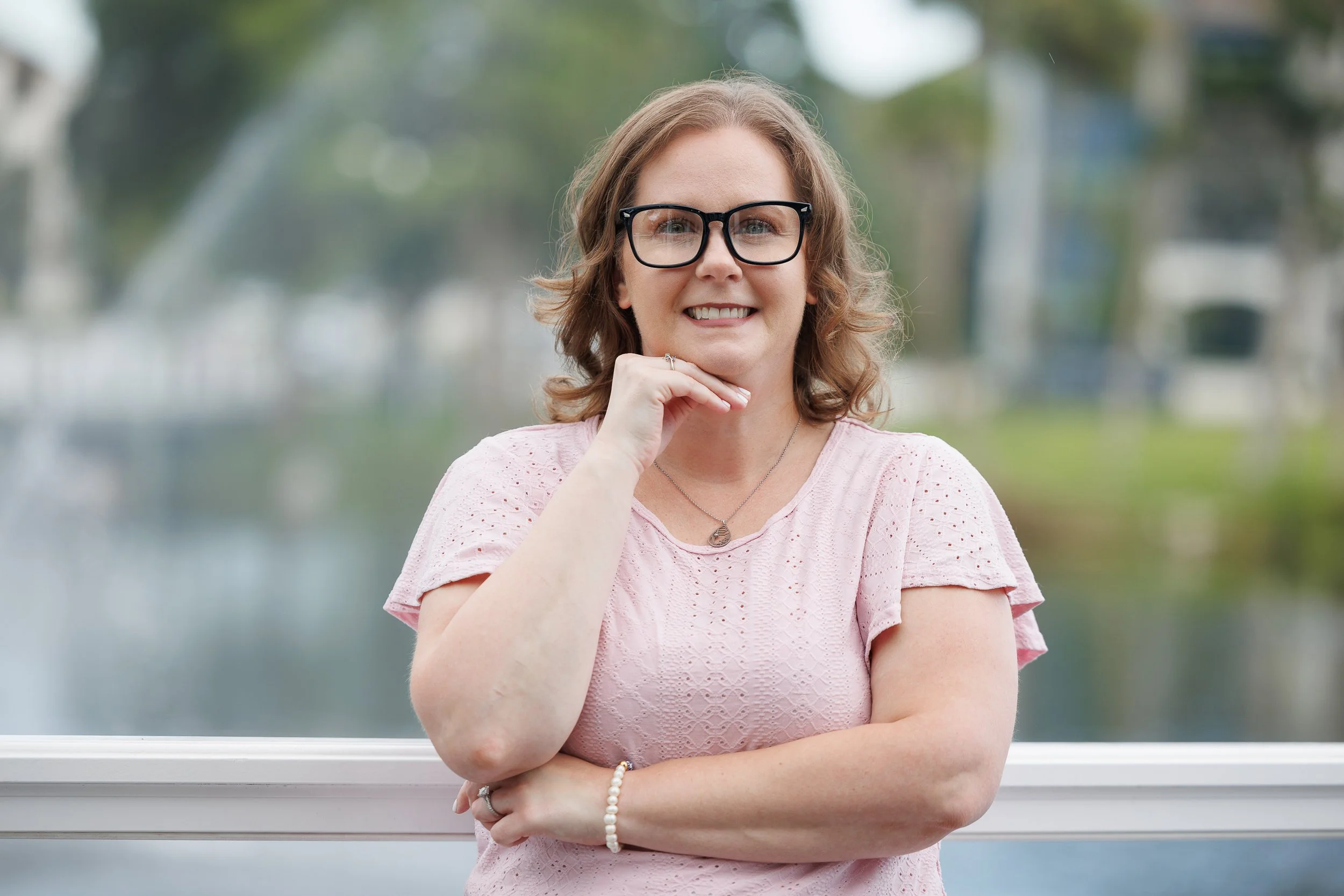 A woman with glasses smiling outdoors, wearing a pink top, with her hand resting on her chin.