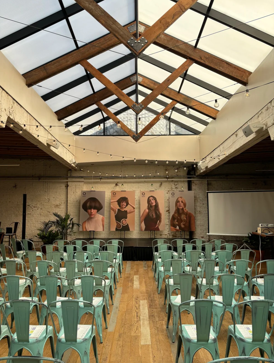 image of a skylight with natural light, blue chairs, and a stage with hair models, a projector screen and some plants at the joinery