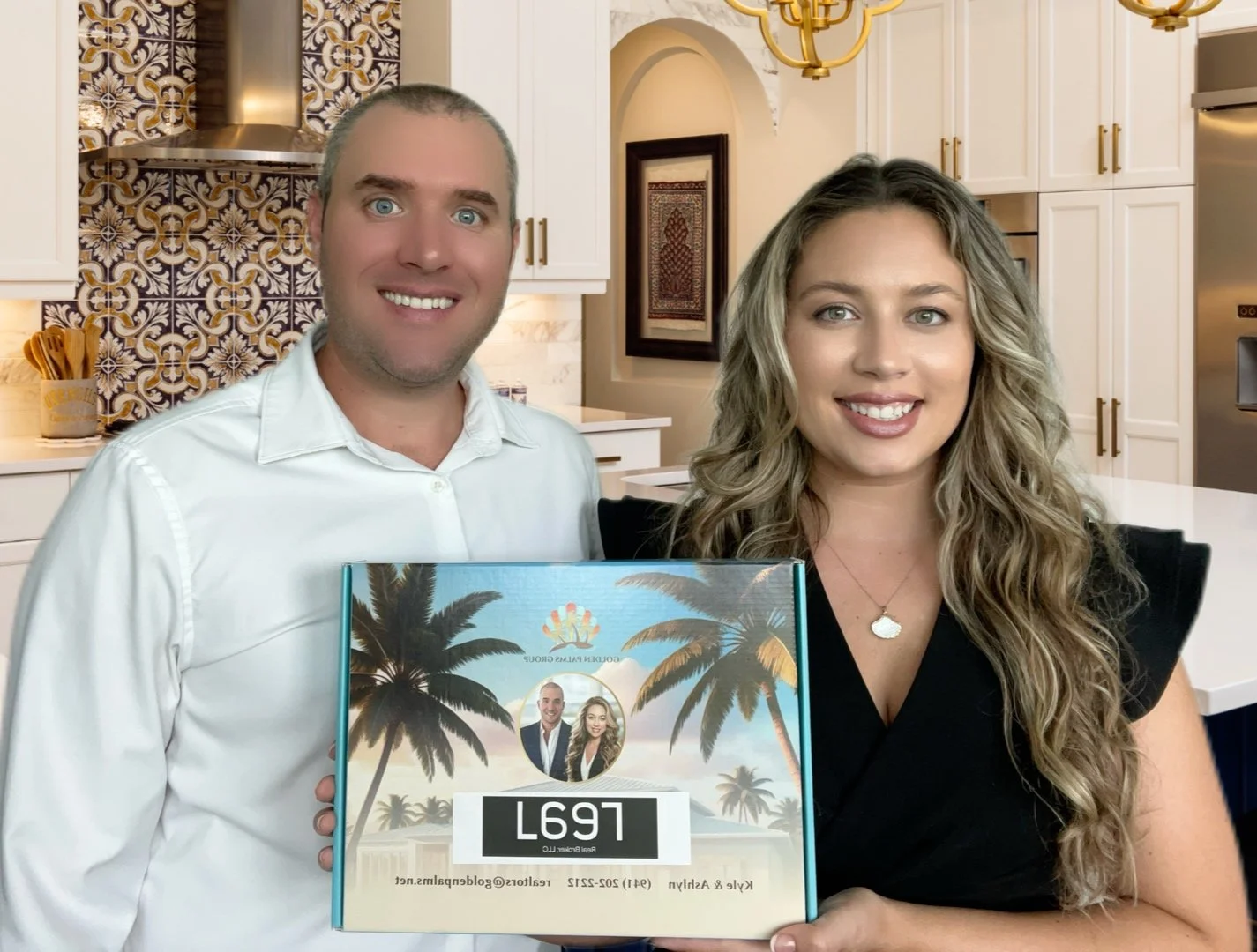 A man and woman smiling and holding a book or journal with palm trees and a small circular photo of themselves on the cover, in a modern kitchen.