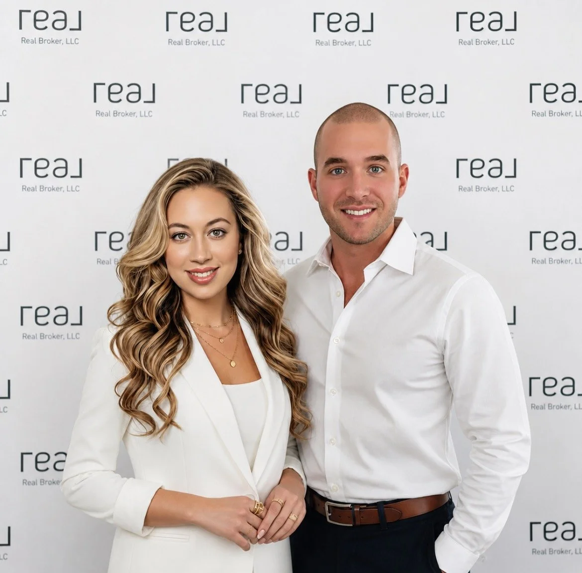 A man and a woman in business attire standing in front of a step and repeat backdrop with the logo 'Real' and the text 'Real Broker, LLC' repeatedly printed on it, smiling at the camera.