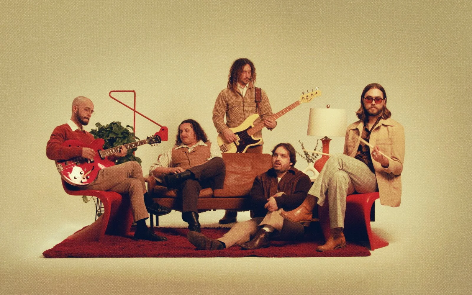 Five individuals in vintage clothing with musical instruments in a room with minimalist decor, including a lamp, a red rug, and a plant.