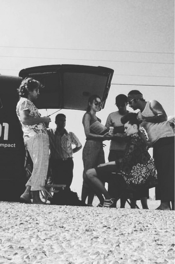 Group of people, primarily women, standing and sitting outdoors near a bus, engaged in conversation and reading documents, with some wearing vintage clothing.