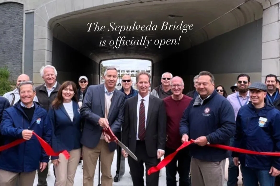 A group of people at the opening ceremony of the Sepulveda Bridge, with a man cutting a red ribbon. The background shows an underpass and a boat passing under the bridge.