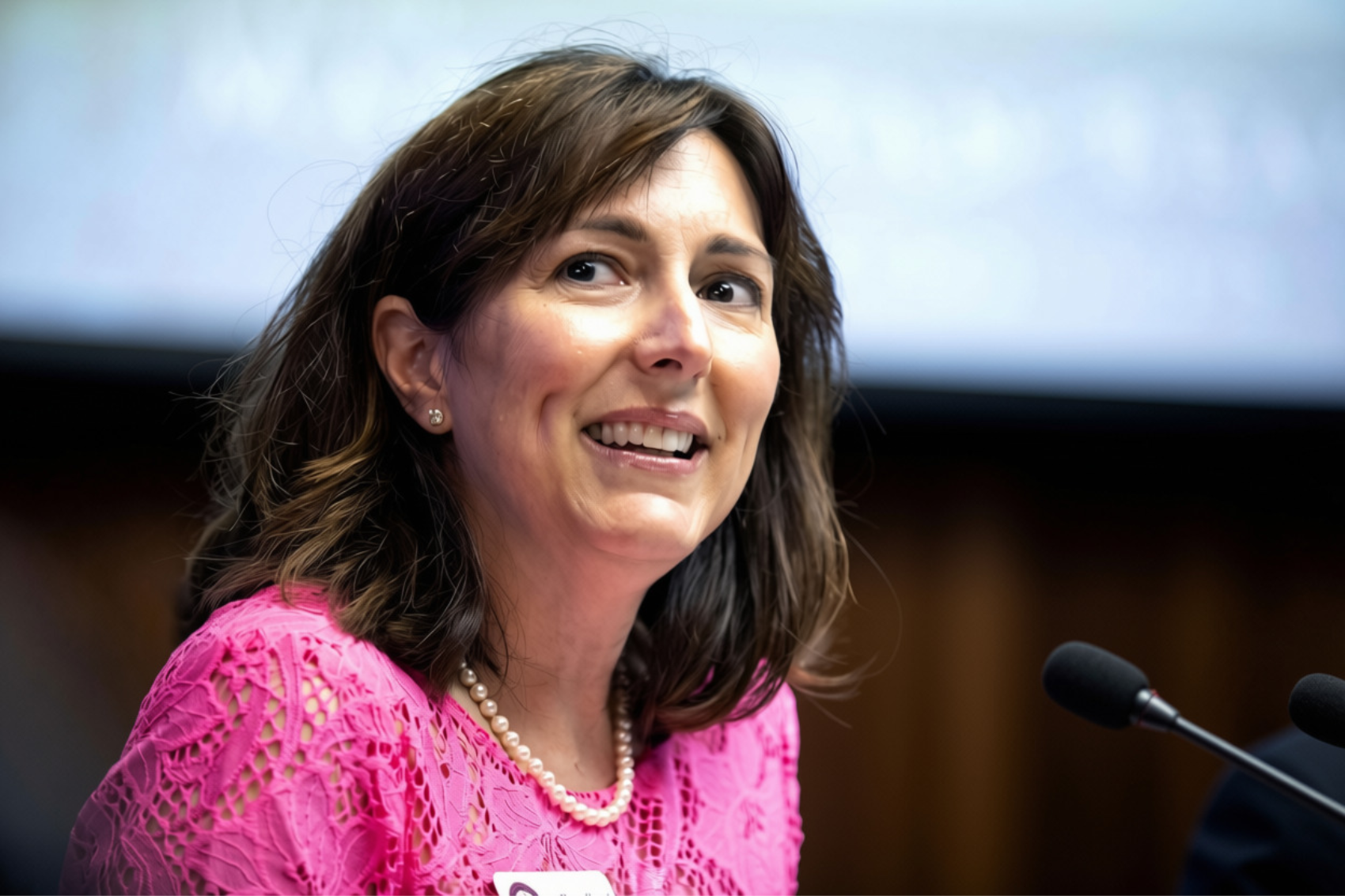 A woman with shoulder-length brown hair wearing a pink lace dress and pearl necklace speaking at a microphone.
