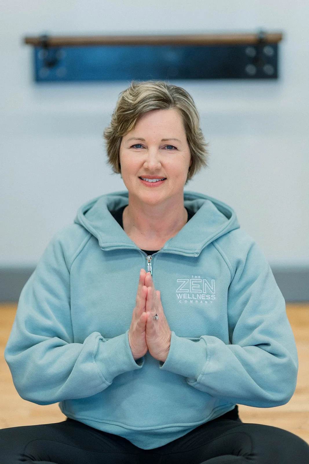 A woman with short gray hair practicing yoga or meditation, sitting cross-legged with hands pressed together in front of her chest, wearing a light blue hoodie with 'The Zen Wellness Company' logo, in a wellness studio.