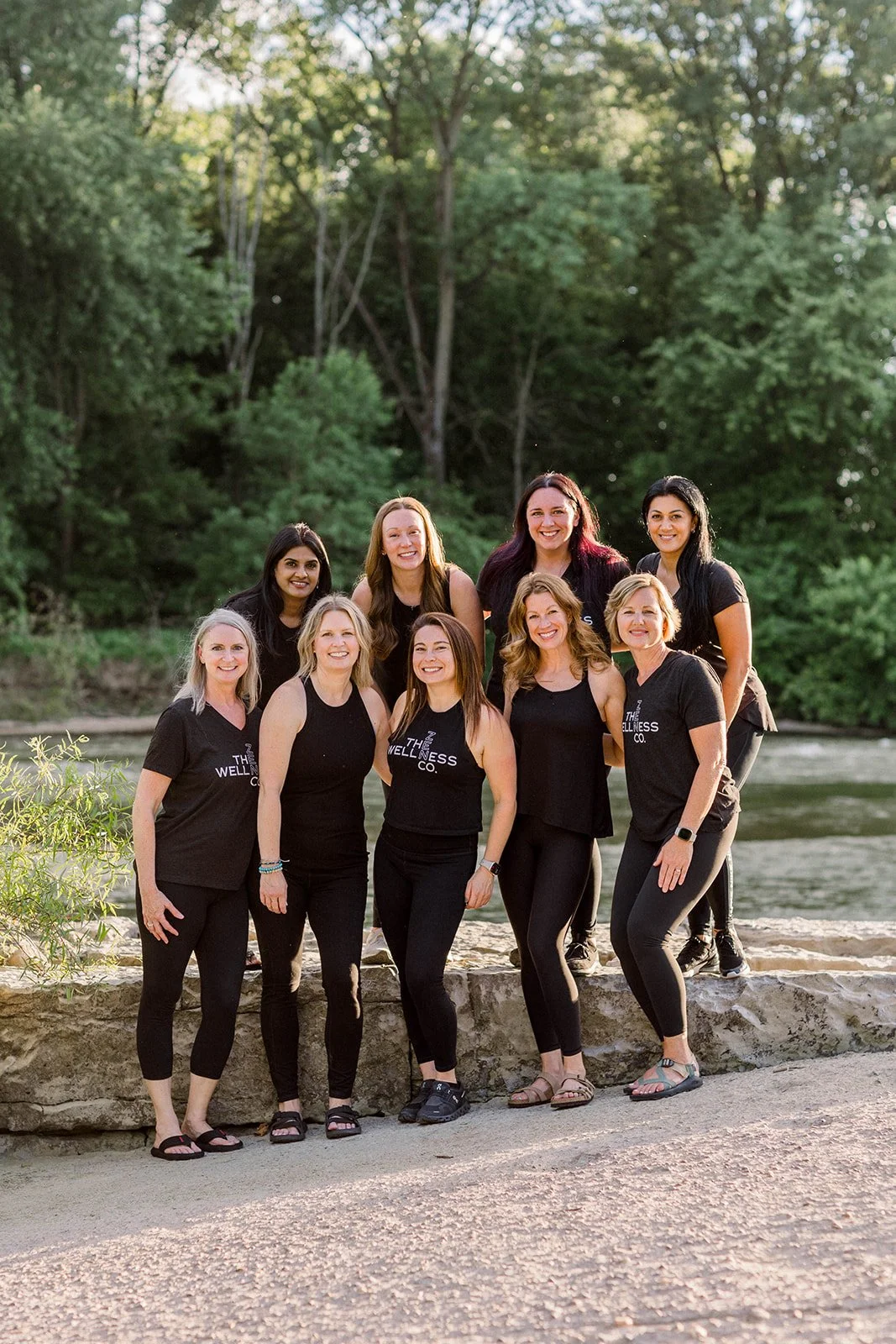 A group of nine women standing on a stone ledge near a river, outdoors with trees in the background, smiling for the camera, some wearing black tops with 'The Wellness Co.' written on them.