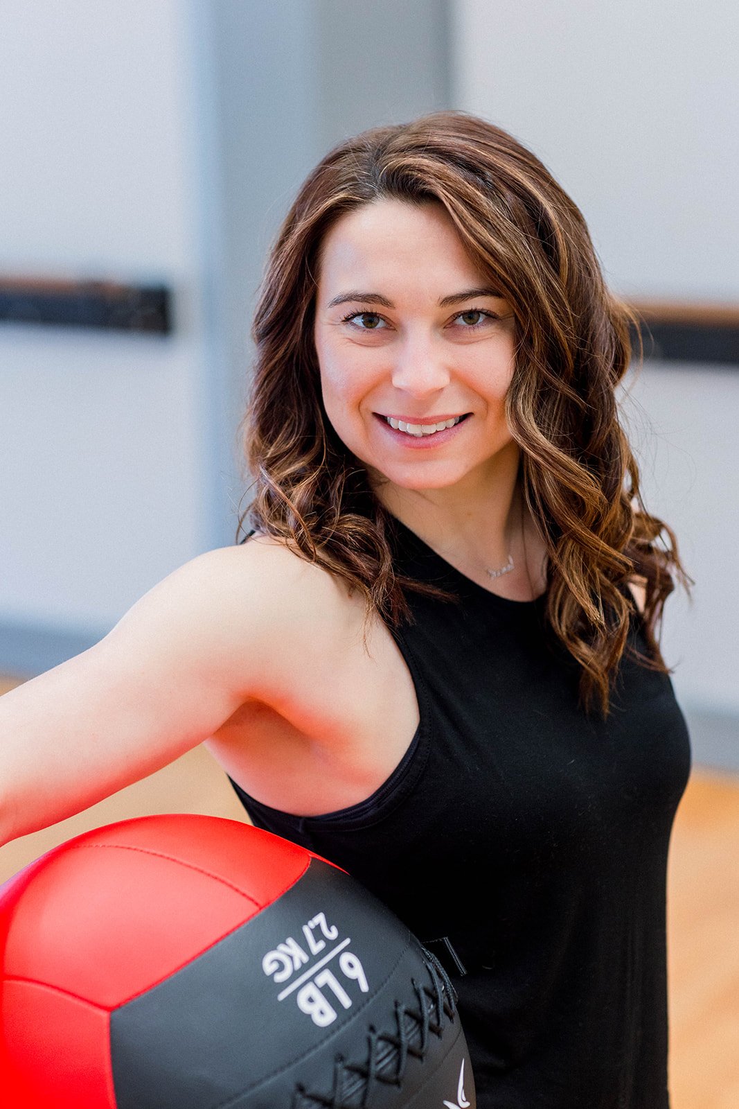 A smiling woman with brown wavy hair wearing a black tank top, holding a red and black exercise ball with the text 'PLB 22KG' on it, in an indoor fitness setting.