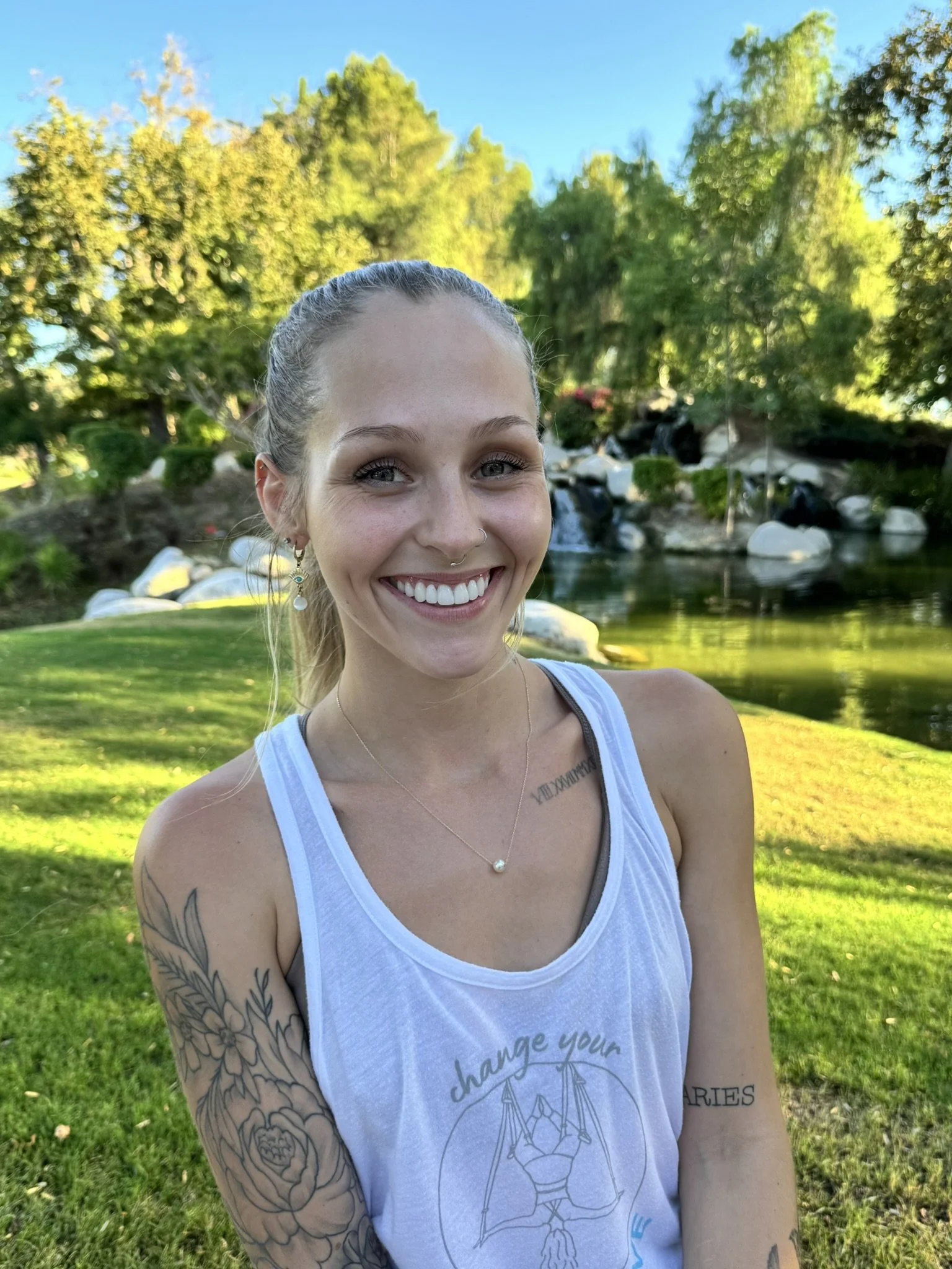 Young woman smiling outdoors with trees, rocks, and a pond in the background during daytime.