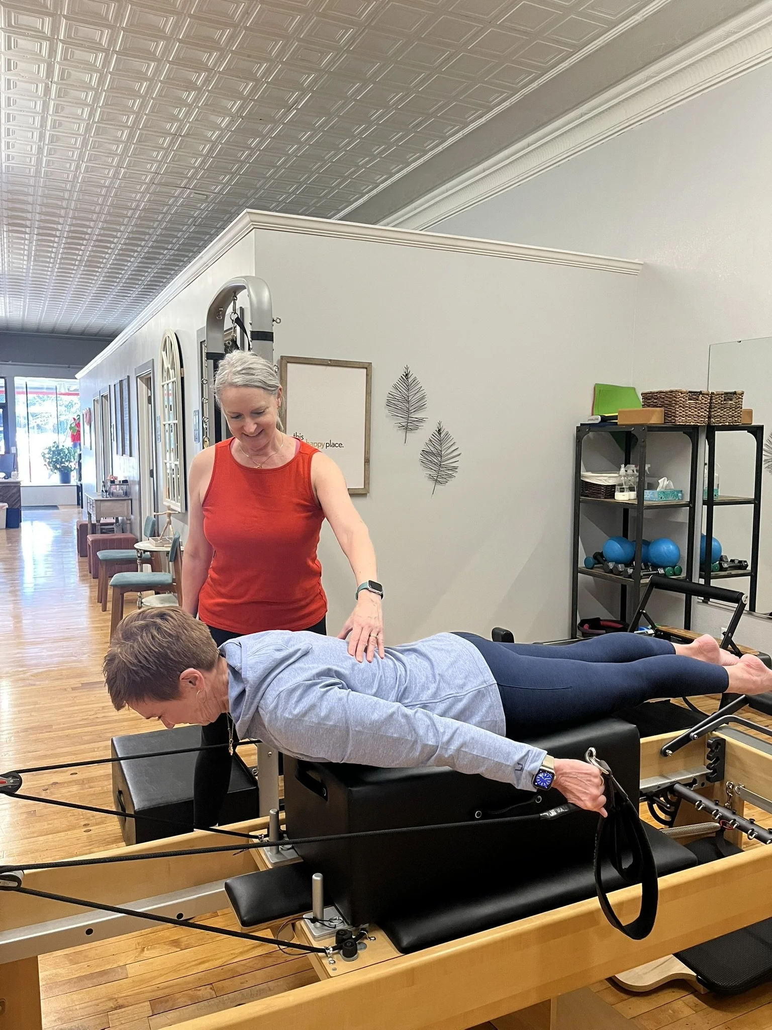 A woman wearing a red top is performing chiropractic or physical therapy on a patient lying face down on a therapy table. The room has wooden floors, a decorative ceiling, and nearby shelves with exercise equipment.