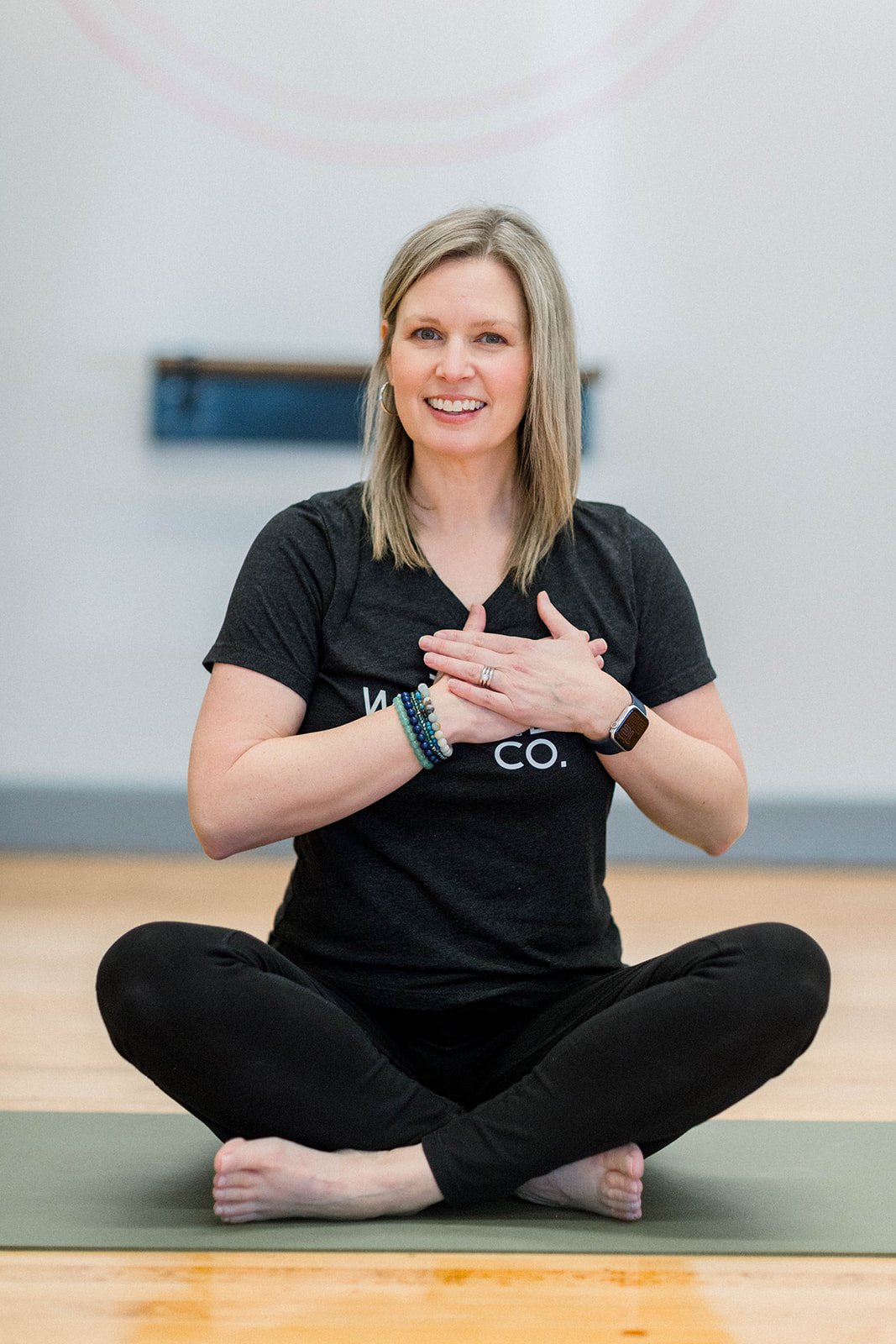A woman sitting cross-legged on a yoga mat with her hands over her heart, smiling and looking at the camera.
