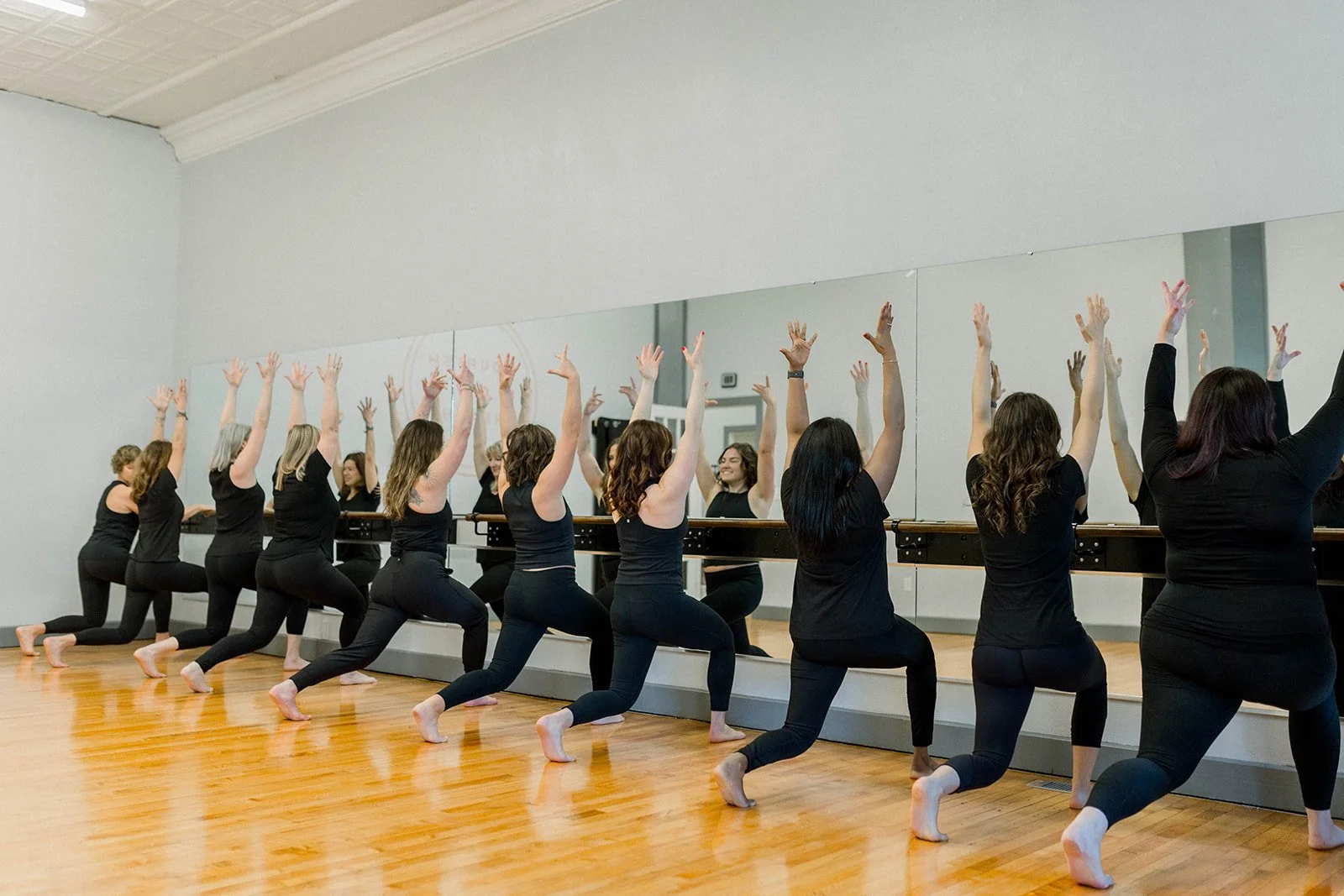 Women in black workout attire practicing ballet at a barre in front of a mirror in a dance studio.