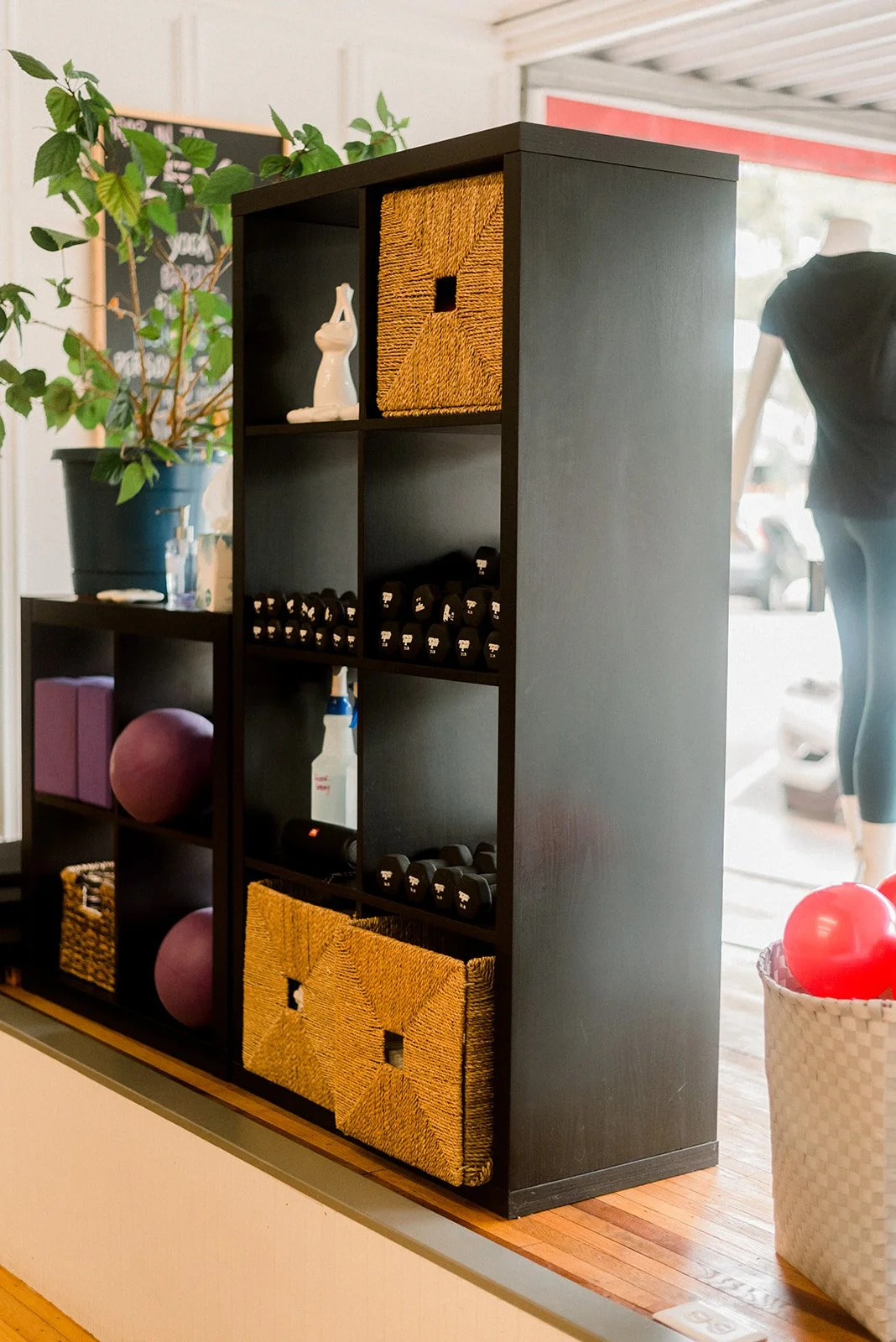 Black shelving unit with storage baskets, dumbbells, exercise ball, and decorative items in a gym or wellness space.