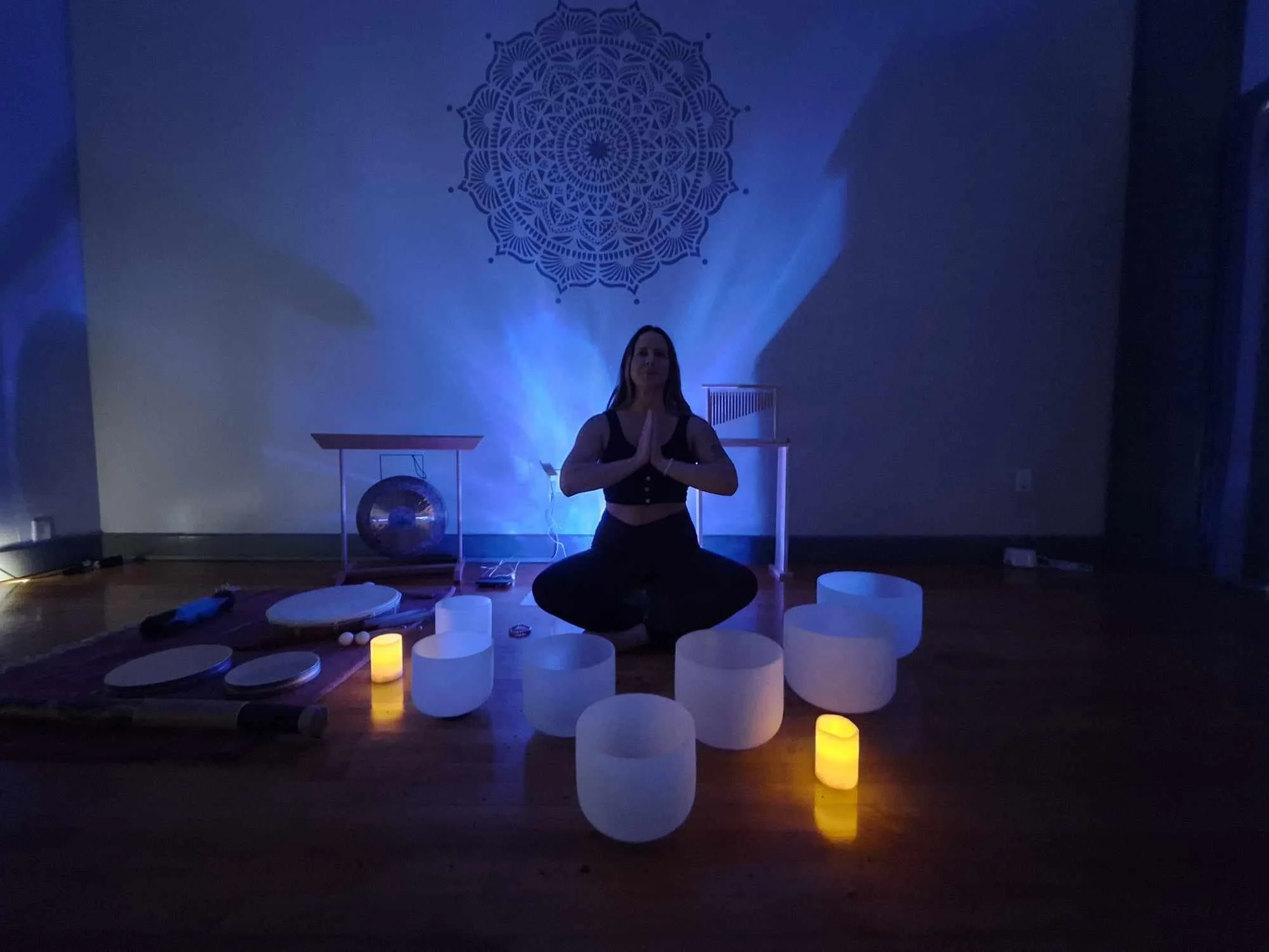 A woman practicing yoga in a dimly lit room with blue lighting. She is kneeling with her hands pressed together in a prayer position. The room is decorated with a mandala pattern on the wall, illuminated by the blue light. Candles are placed around her on the floor, creating a peaceful atmosphere.
