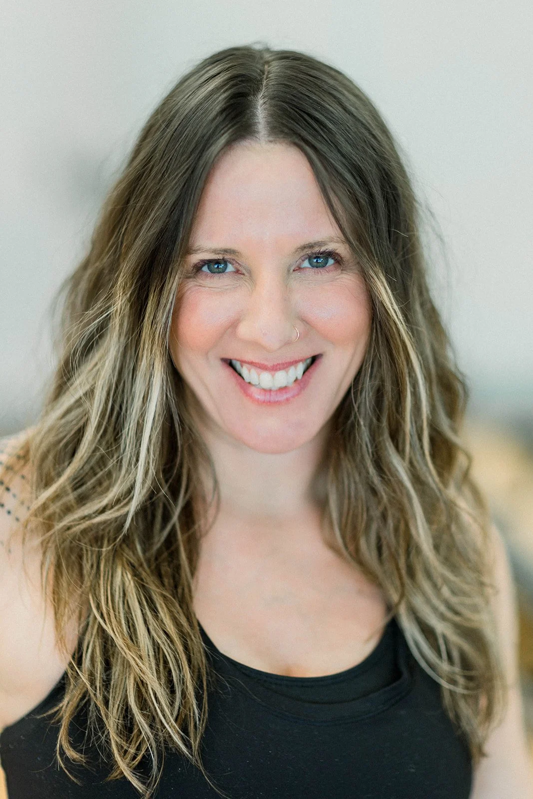 A woman with long wavy brown hair, blue eyes, and a nose ring smiling at the camera. She is wearing a black sleeveless top.
