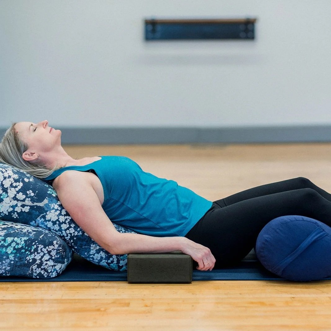 Woman lying on a yoga mat with bolster and blocks, practicing restorative yoga in a studio.