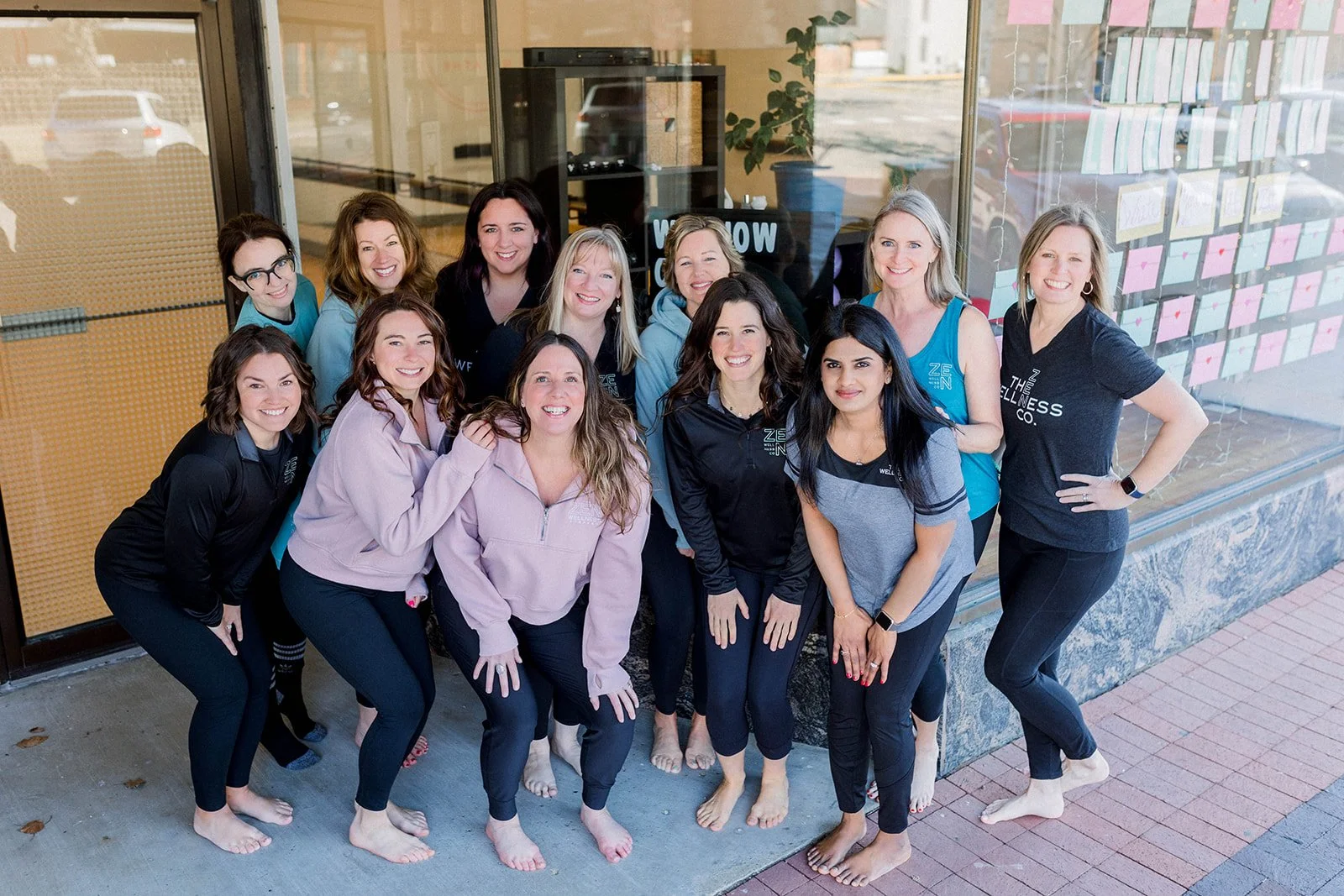 A group of fifteen women standing outside a building, smiling and posing for a photo. Some women are barefoot, and some are wearing matching shirts with a wellness-related logo.