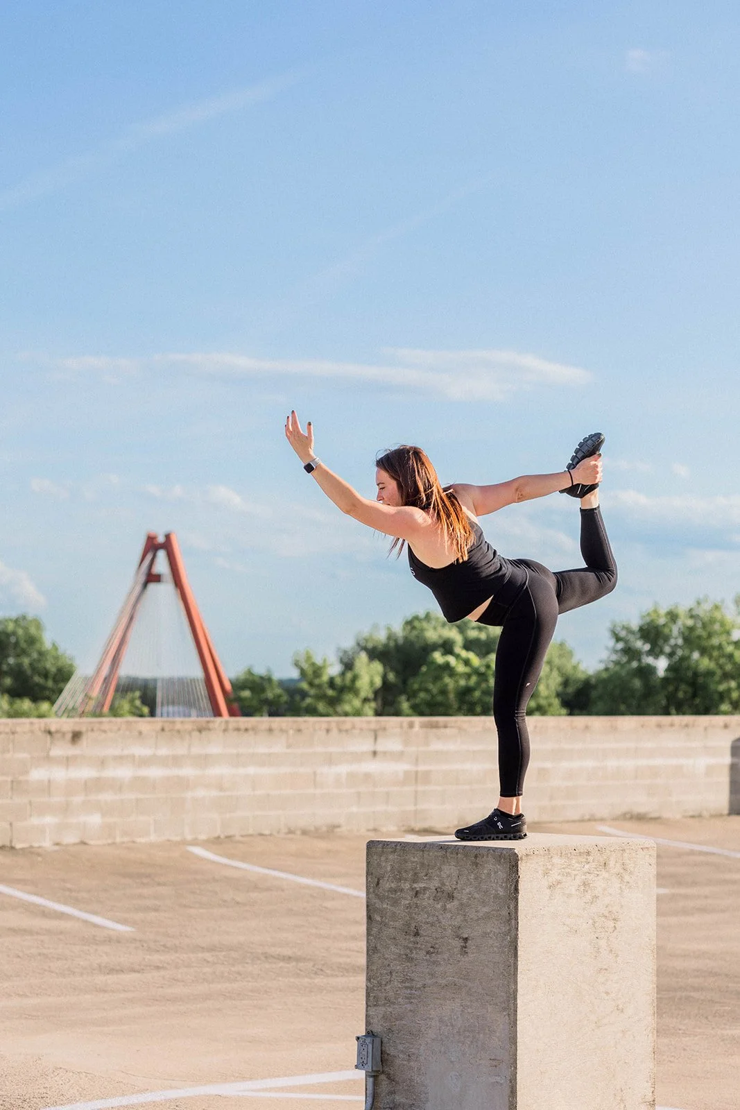 Woman performing a yoga pose on a concrete block in an outdoor parking lot with a bridge and trees in the background on a sunny day.