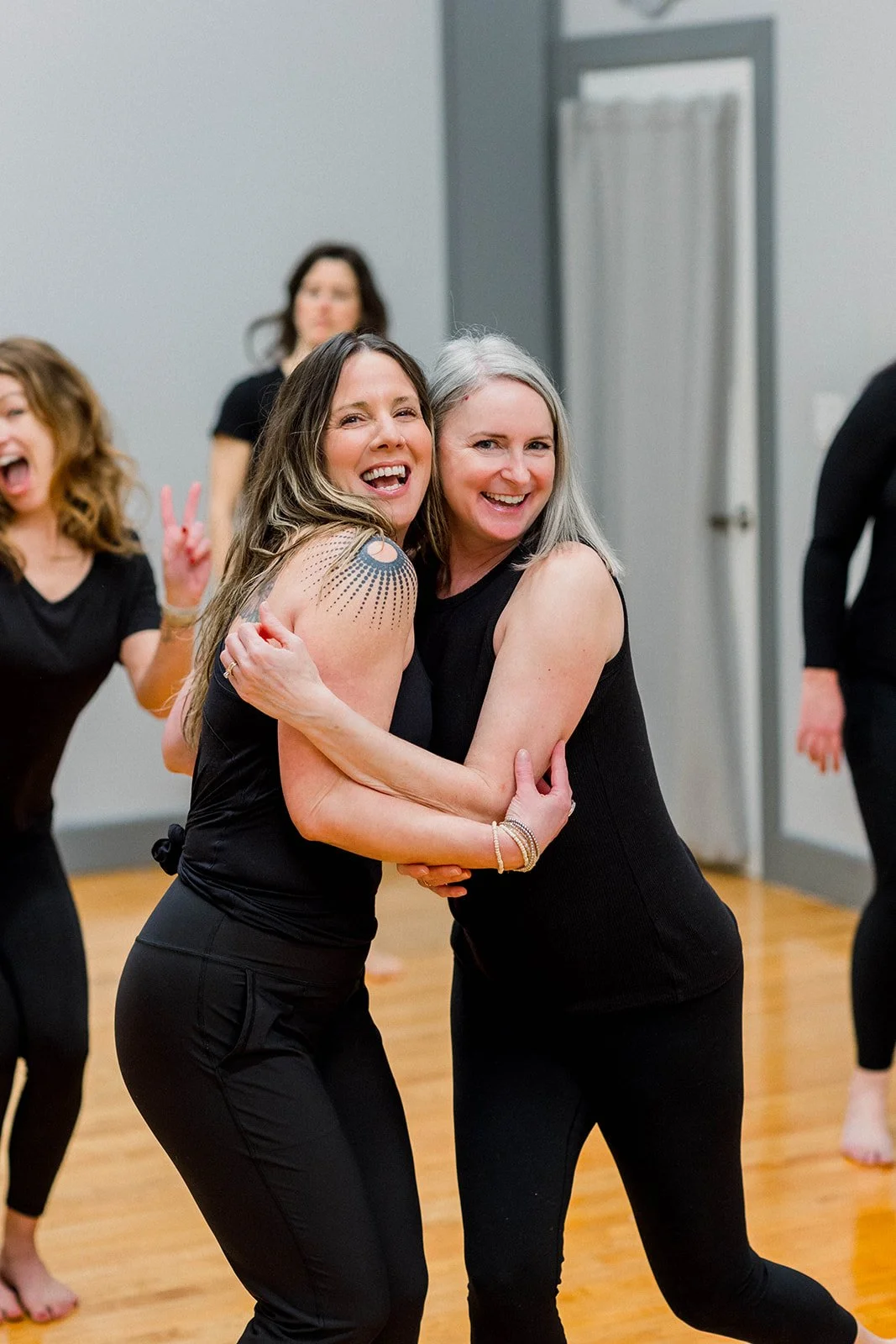 Two women embracing and smiling, dancing at a social gathering, with other women in the background making playful gestures.