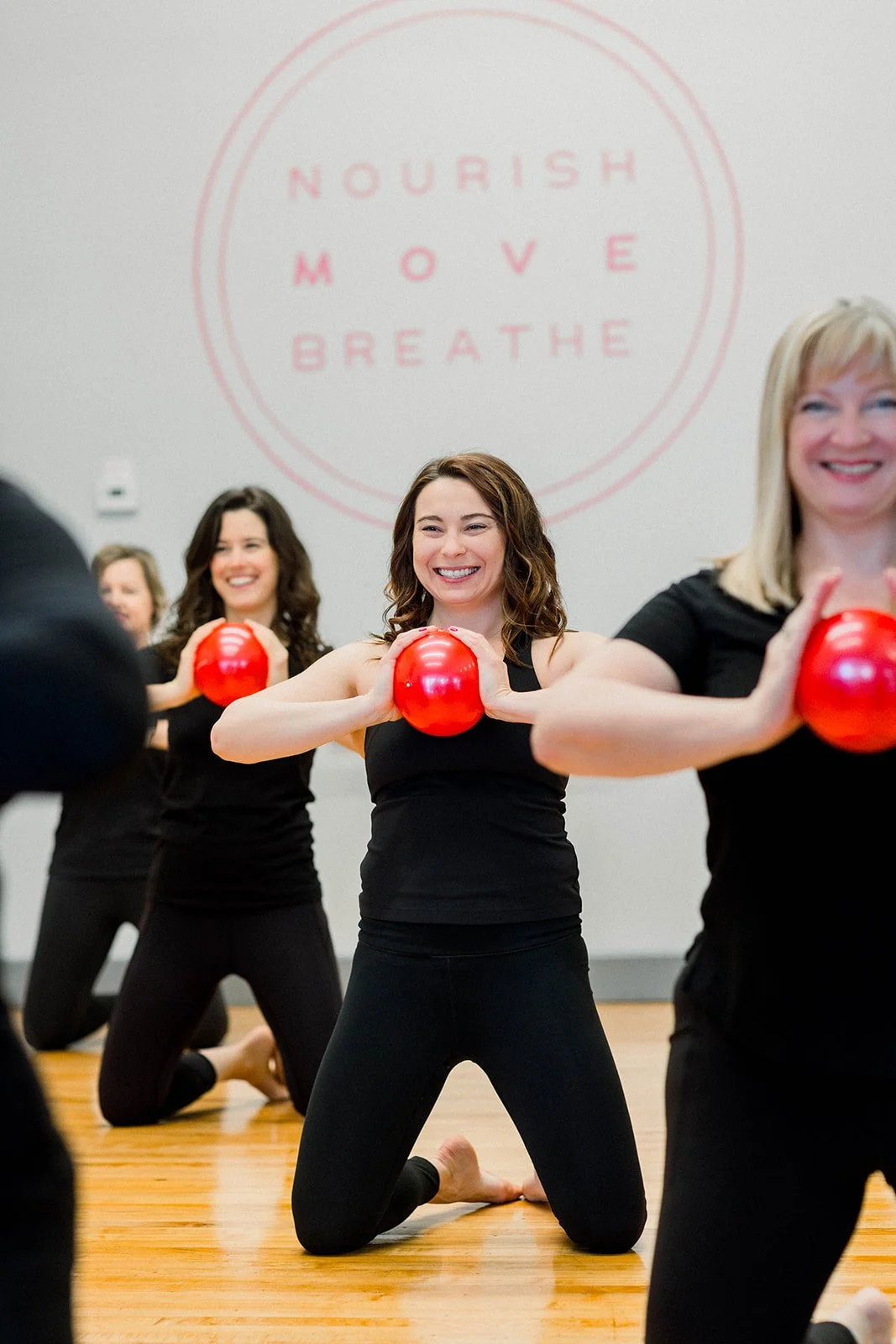 Group of women practicing a fitness class with red exercise balls, kneeling on a wooden floor in a studio with a motivational sign on the wall that reads 'Nourish, Move, Breathe'.
