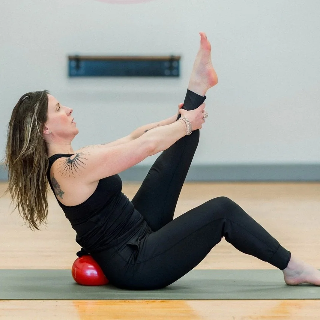 Woman practicing yoga seated on a mat with a small red ball under her lower back, stretching her legs and reaching for her foot.