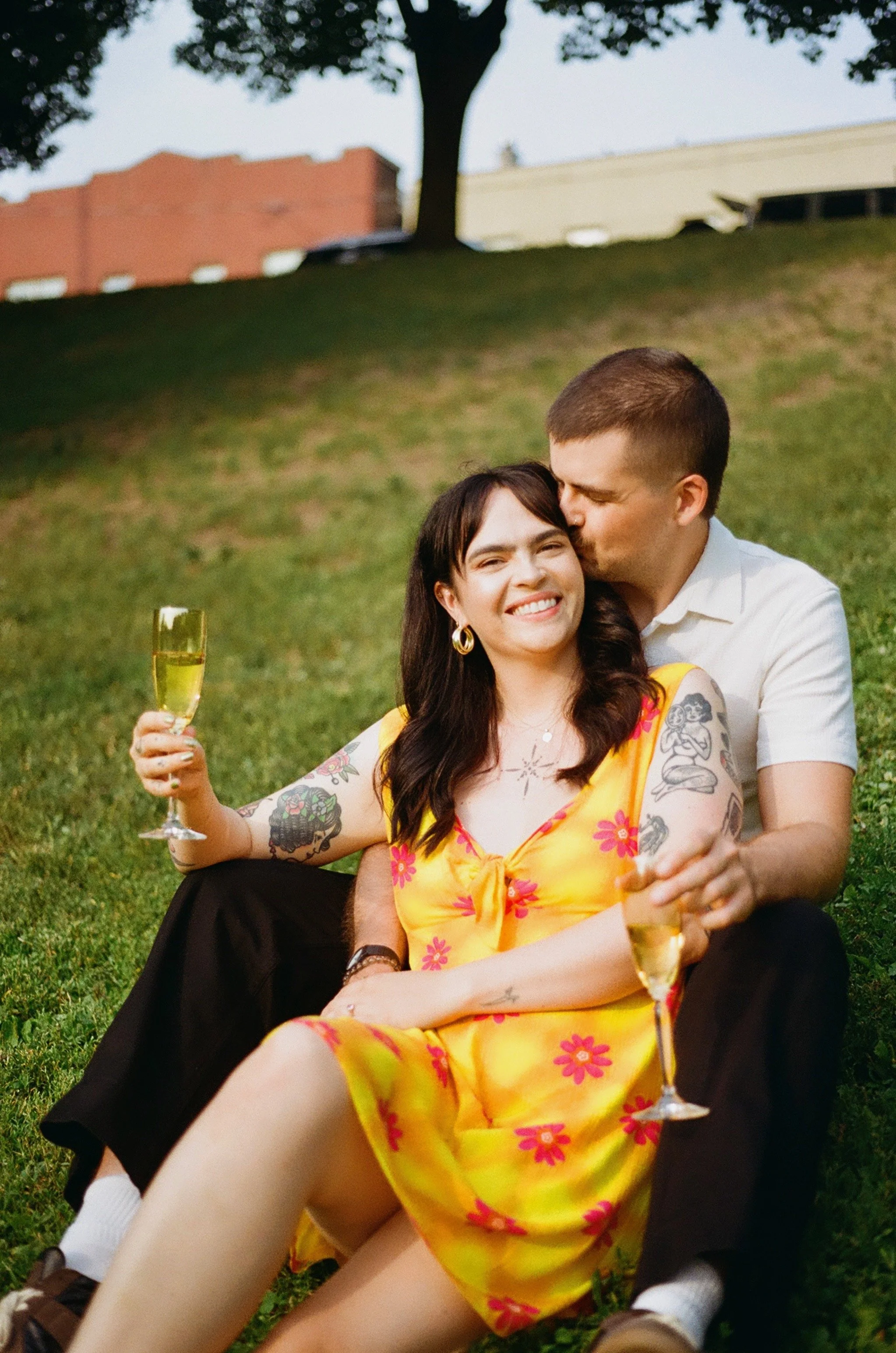 A man kissing a woman on the cheek while sitting on grass. The woman is smiling and holding a champagne glass. They are outdoors with trees and buildings in the background, enjoying a casual moment together.
