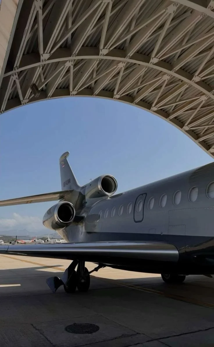 View of a private jet airplane parked at an airport hangar, with part of the hangar's roof visible overhead.