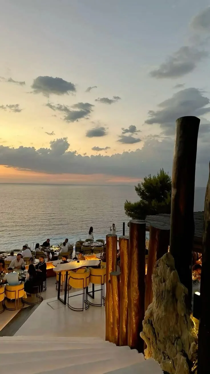 A beachside restaurant at sunset with tables and chairs, some patrons dining, overlooking the ocean with a partly cloudy sky.