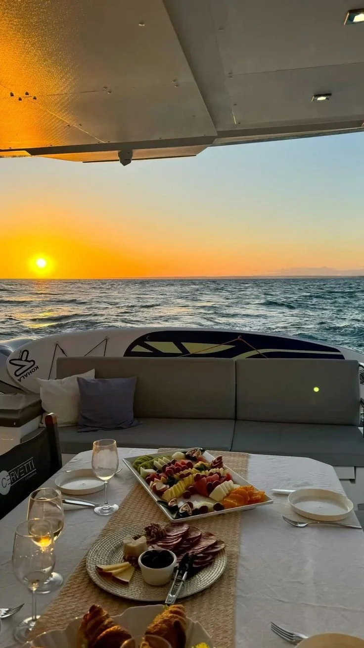A dining setup on a boat with a charcuterie and fruit platter, glasses of wine or champagne, and a sunset over the ocean in the background.