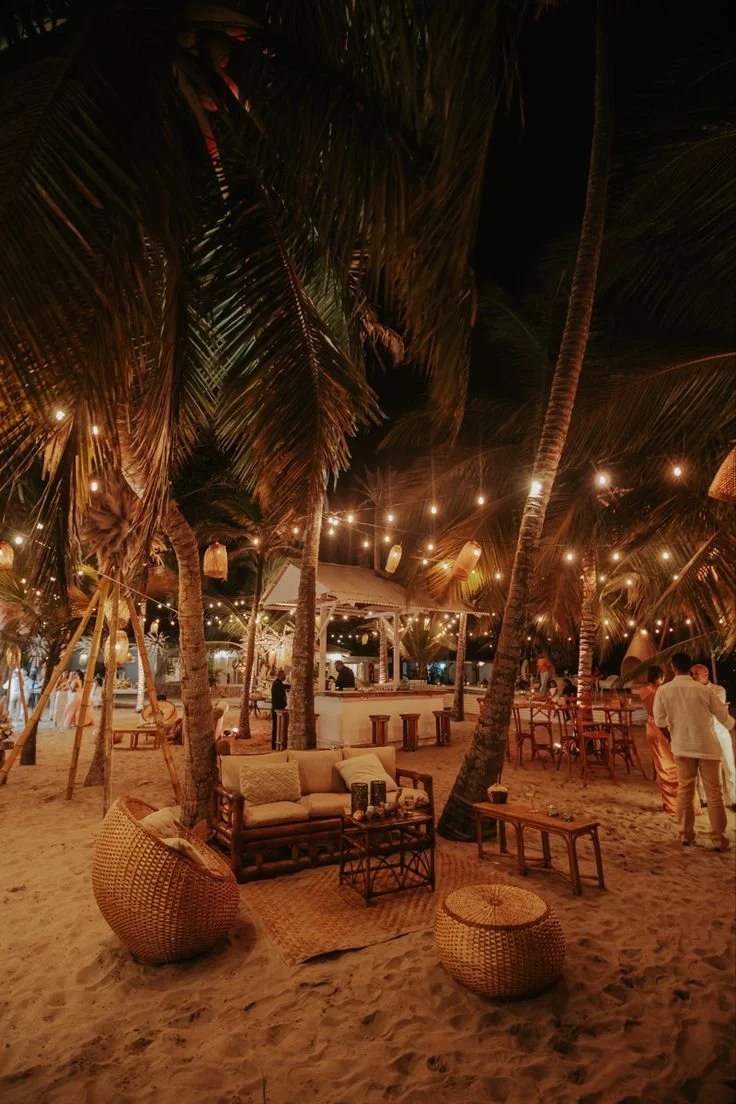 Beachside lounge area at night with hanging lanterns and string lights, wicker furniture, palm trees, and people socializing.