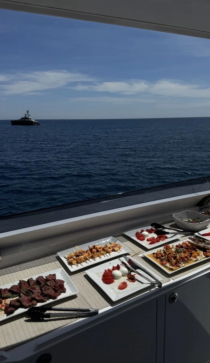 Assorted appetizers on plates, including sliced beef, skewered shrimp, and vegetable dishes, set on a table with a view of the ocean and a boat in the distance.